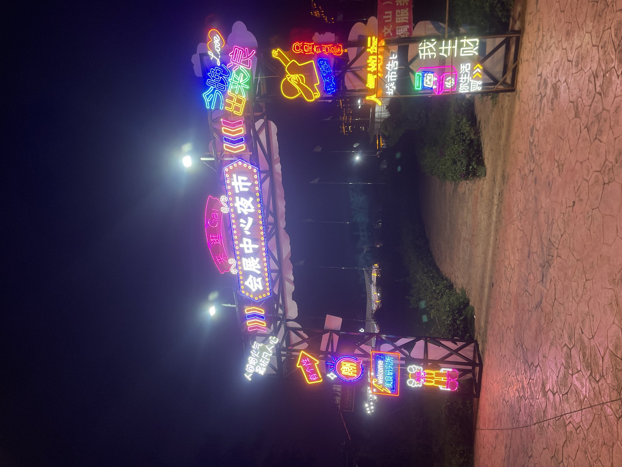 An eye-level, night shot captures a brightly lit archway adorned with neon signs, marking the entrance to a night market in an unknown city. The archway features a prominent neon sign in Chinese characters that translates to "Convention Center Night Market." Flanking the central sign are other neon decorations, including a pair of hands in a dabbing pose, a cartoonish figure, and text in English and Chinese that reads "welcome" and "Gourmet Research Institute." The ground in front of the archway is paved with large, irregularly shaped stones that reflect the neon lights. To the right of the archway, a vertical neon sign in Chinese characters says "Popularity" and "City Announcement." Further to the right, another neon sign displays the Chinese characters for "I," "Life," and "Wealth," along with shopping cart icons. Beyond the archway, the night market appears to be set up with tents and stalls, illuminated by various lights, suggesting a bustling commercial and social gathering. The overall scene is a vibrant display of nocturnal commerce and entertainment, typical of a city's night market offerings.