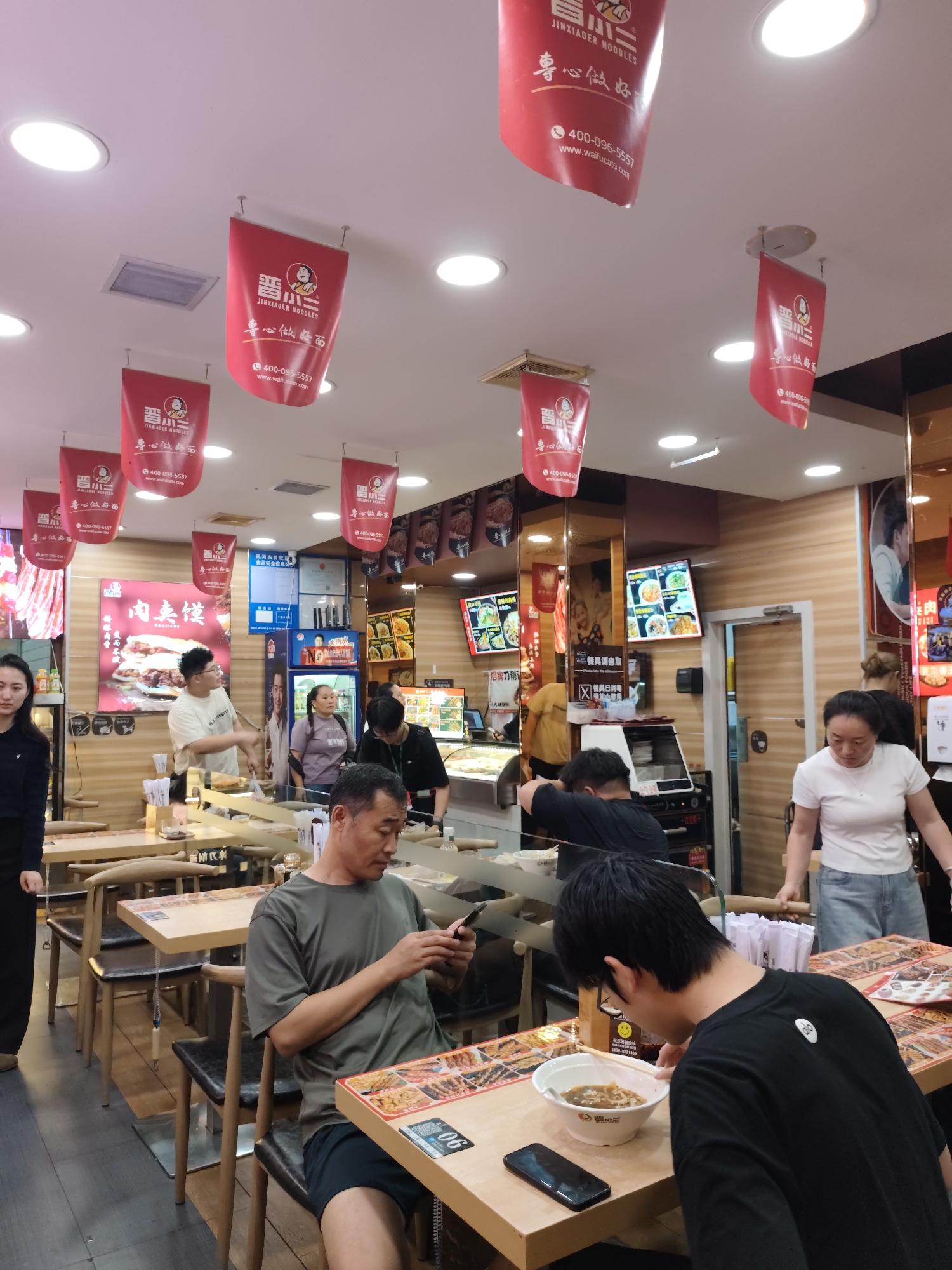The image depicts the interior of a restaurant, likely a noodle shop, with red banners hanging from the ceiling displaying the brand "JINXIADER NOODLES" and Chinese characters. Several customers are present. In the foreground, a man in a green t-shirt and black shorts is seated at a table, looking at his phone. To his right, another person, mostly out of frame, is eating noodles. A young person in a black t-shirt is also seated at a table, eating from a bowl of noodles. Further back, a woman in a white t-shirt and denim shorts stands near a counter, and other staff members are visible behind the counter. The restaurant appears to have wooden tables and chairs, with bright overhead lighting. The overall ambiance suggests a casual dining environment. The presence of specific branding and the style of the establishment imply it is a chain restaurant. It is noted that this image was captured in Aihui, China.