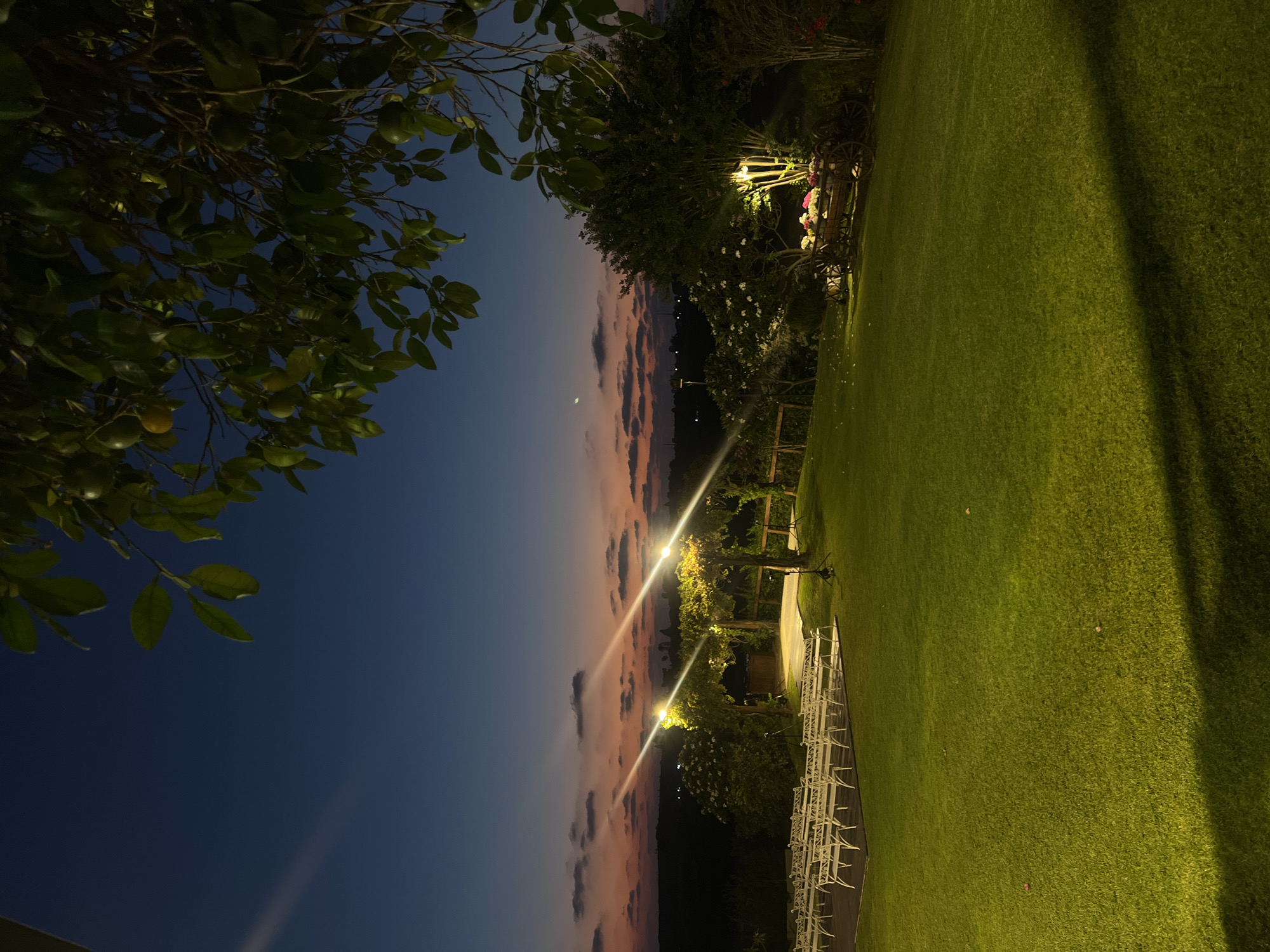This is a photograph taken in Beit Hanan, Israel, at dusk. The sky displays a gradient of deep blue at the top, transitioning to hues of orange and pink near the horizon, with scattered clouds. The foreground is dominated by a well-maintained, brightly lit green lawn. To the left, rows of white chairs are arranged on a wooden platform, suggesting an outdoor event setup.
In the middle ground, a raised platform with a white surface is visible, illuminated by overhead lights. The area around this platform is decorated with trellises and foliage. Further to the right, a tree is lit by a spotlight, highlighting its branches and a rustic wooden cart filled with flowers. The background shows a silhouetted landscape with distant lights. The overall scene is tranquil and suggests a venue prepared for an evening gathering.