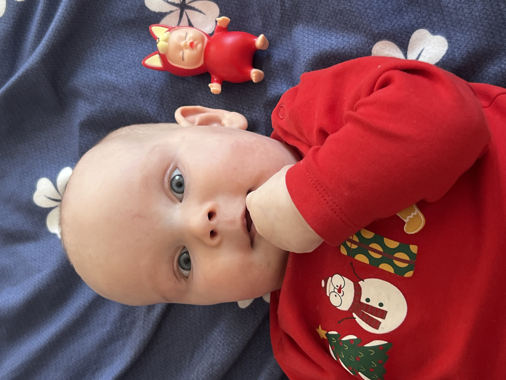 A baby in a red, long-sleeved Christmas-themed onesie lies on a blue floral patterned surface. The baby has light skin, blue eyes, and is looking at the camera with their hand near their mouth. A small red doll with yellow accents is positioned near the baby's head. The backdrop appears to be bedding with white floral designs against a blue background. The location is unknown and cannot be ascertained from the image. The city of Votkinsk, Russia is not identifiable in the image.
