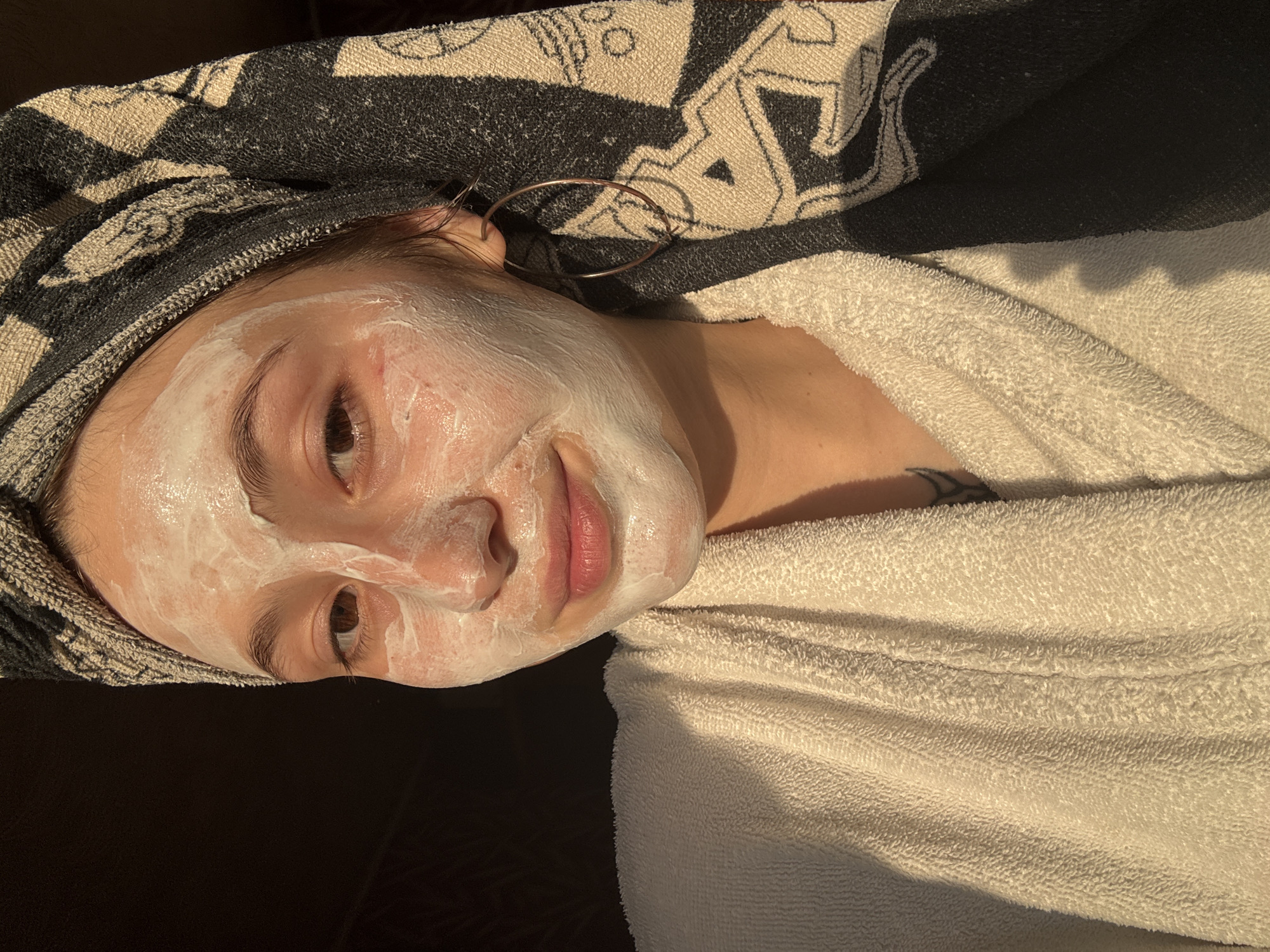 A young adult female is depicted in a medium close-up, facing directly forward. Her face is largely covered by a white, creamy facial mask, applied from the forehead down to the chin, leaving the eyes, nostrils, and lips clear. Her eyes are brown and gaze directly at the viewer. Her lips are a natural pink, slightly parted, with a subtle smile.
She is wearing an off-white or cream-colored terrycloth bathrobe, visible on her chest and shoulders. Her head is wrapped in a black and white patterned terrycloth towel, exhibiting an abstract or geometric design. A large, thin, metallic hoop earring is visible on her right ear (viewer's left). On her left shoulder/collarbone area, a dark, partial tattoo with abstract or curved lines is visible peeking from under the robe.
The scene is illuminated by strong, warm, directional light, consistent with sunlight, originating from the viewer's left. This light highlights the left side of her face, the texture of the white mask, the folds of her robe, and the patterned head towel. The background is uniformly dark and indistinct, offering no discernible features or objects.
Based on the visual information provided, the specific location in the city of Фоки, Russia, cannot be determined. The image portrays an indoor setting with a dark, unidentifiable background.