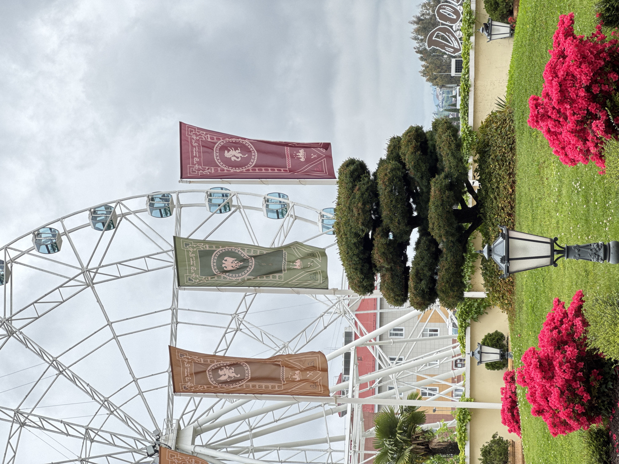 A white Ferris wheel is the focal point of the scene, adorned with maroon, olive green, and brown heraldic banners that display mythical creatures. Behind it are buildings with red and grey roofs. The scene takes place in the Sirius region of Russia. In the foreground, there are evergreen trees and red flowering bushes beside black lampposts on a green lawn. The sky is overcast.