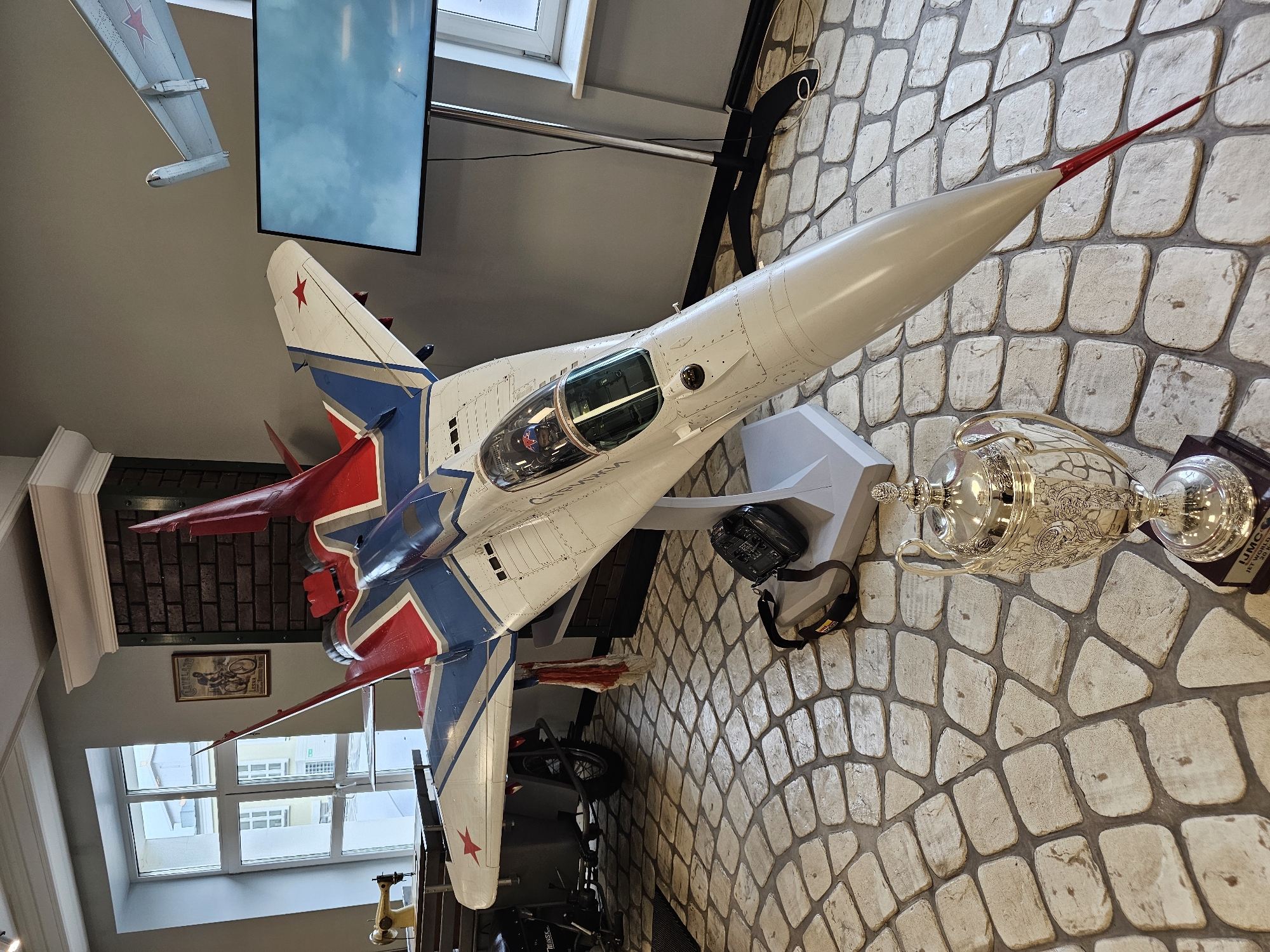A detailed view reveals a room's interior, seemingly a space within a museum, given the presence of a model fighter jet airplane of the MiG-29 Fulcrum variant, adorned with Soviet-era markings and prominently displayed along with a trophy. The model is affixed to a mount against a patterned, cobblestone-like textured wall.
The setting includes visible windows, a television screen, and additional, less distinct objects in the background, suggesting a curated exhibit. The overall lighting is bright, contributing to clear visibility of the artifacts. The location is assumed to be Архангельское, Russia, as per the prompt.