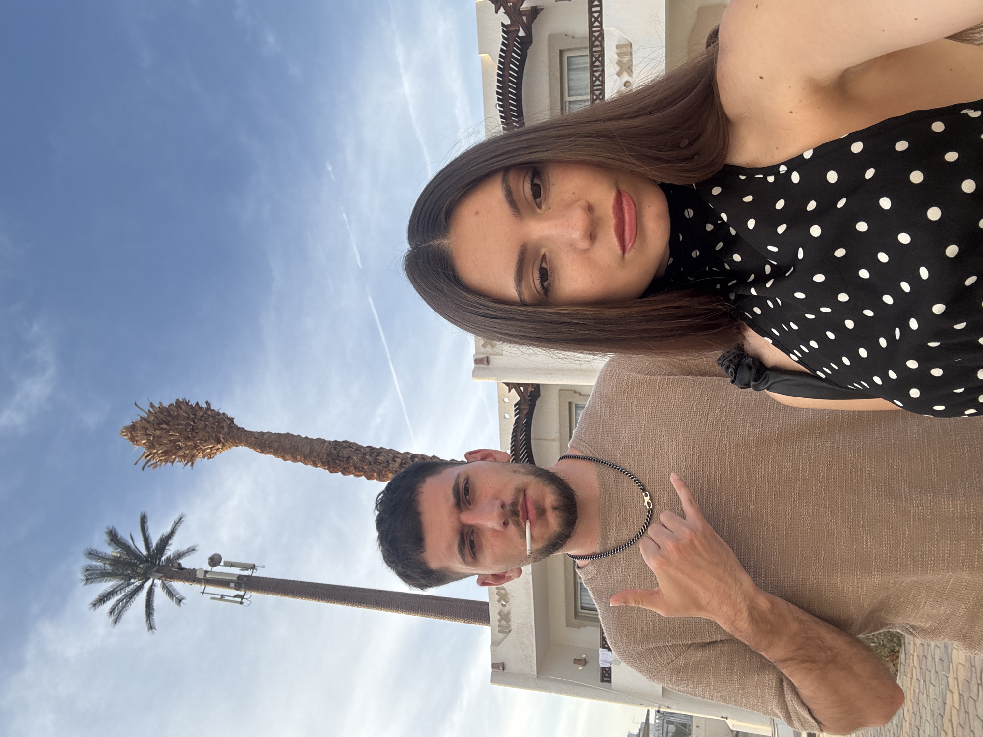 A medium-shot, off-kilter, outdoor photograph taken in Unknown, Unknown, features a light-skinned woman and man. The woman is positioned towards the top of the frame, and the man is near the bottom. She has medium-length brown hair, light-toned skin, and is wearing a black top with white polka dots. The man has short dark hair, a beard, and is wearing a tan sweater. He has a cigarette in his mouth and is gesturing with his right hand. Behind them is a white building with decorative architectural elements. To the left, there are two palm trees against a blue sky with wispy clouds. One of the palm trees appears to have communication equipment attached to it. The image appears to be a selfie or taken at an unusual angle.
