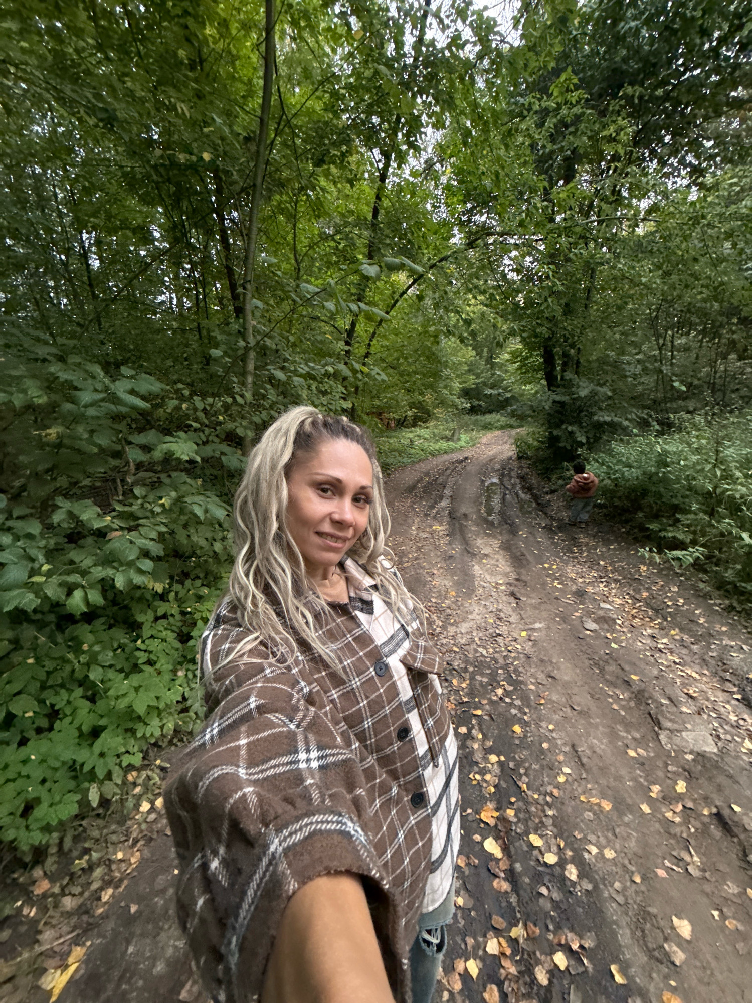 A full-frame, eye-level selfie taken in a forest on a dirt road. The foreground features a woman with blonde hair in a high ponytail, wearing a brown and white plaid shirt. She is smiling at the camera and holding the phone out to take the picture. In the background, a young child in a brown shirt and grey pants is walking away from the camera down the muddy, leaf-strewn road. The road is flanked by dense green trees and foliage. The sky is overcast. The location is identified as Rusanovo, Russia.