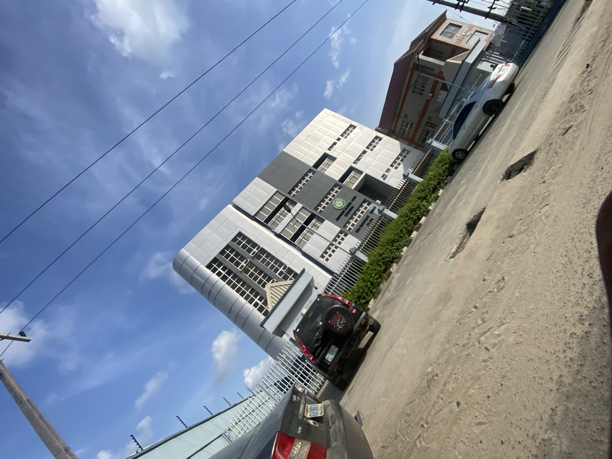 The image shows a building with a sign indicating it is the "Ikorodu National Industrial Court" in Ikorodu, Nigeria. The building has a modern design with multiple windows and a gray and white color scheme. A fence runs along the front of the property. Several cars are parked in front of the building. The sky is blue with scattered clouds, and power lines are visible in the upper-left corner. The foreground shows a paved surface with visible imperfections.