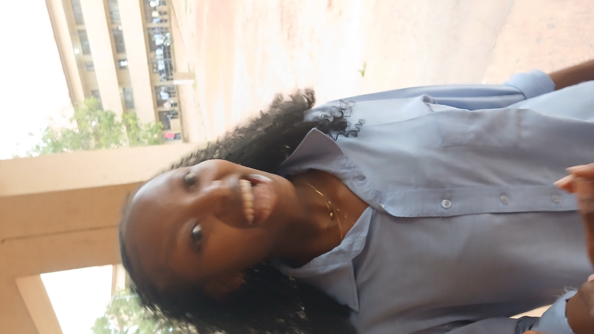 A young woman with dark curly hair, wearing a light blue button-down shirt and a gold necklace, is smiling and looking at the camera. The background shows a building and some trees, suggesting an outdoor location in Jos, Nigeria.