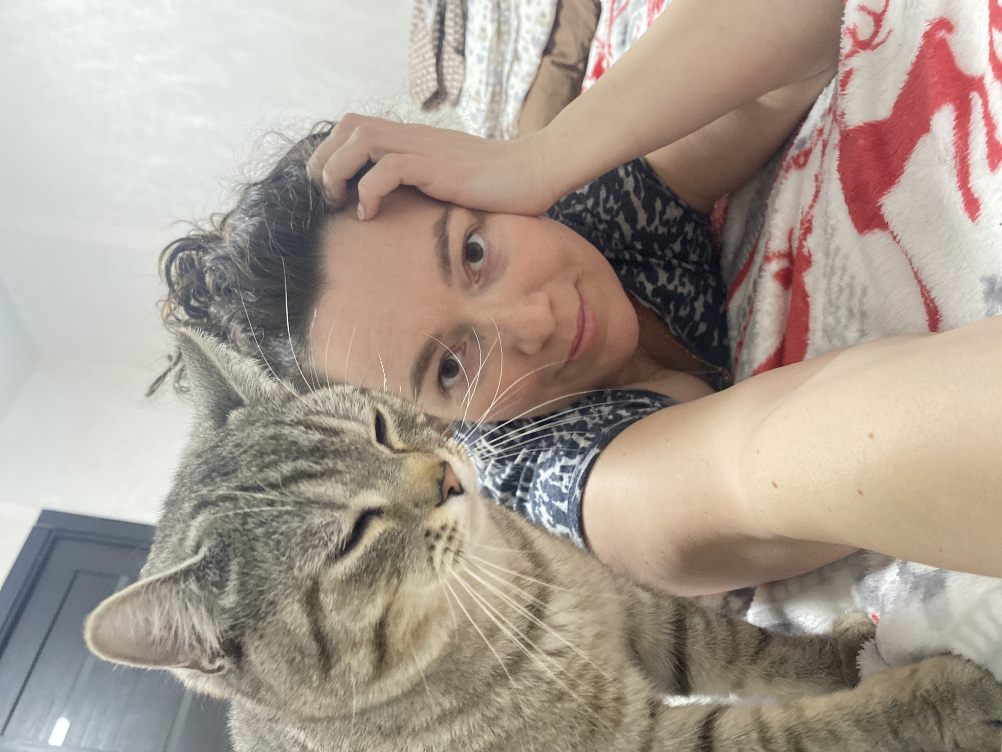 A woman lies on a bed with a grey tabby cat. She rests her head on her hand and smiles at the camera. She is wearing a dark blue and white patterned top. The cat is positioned in the foreground, partially obscuring her arm. The bed is covered with a red and white blanket. A door is visible in the background. The location is in the city of Ольховка, Russia.