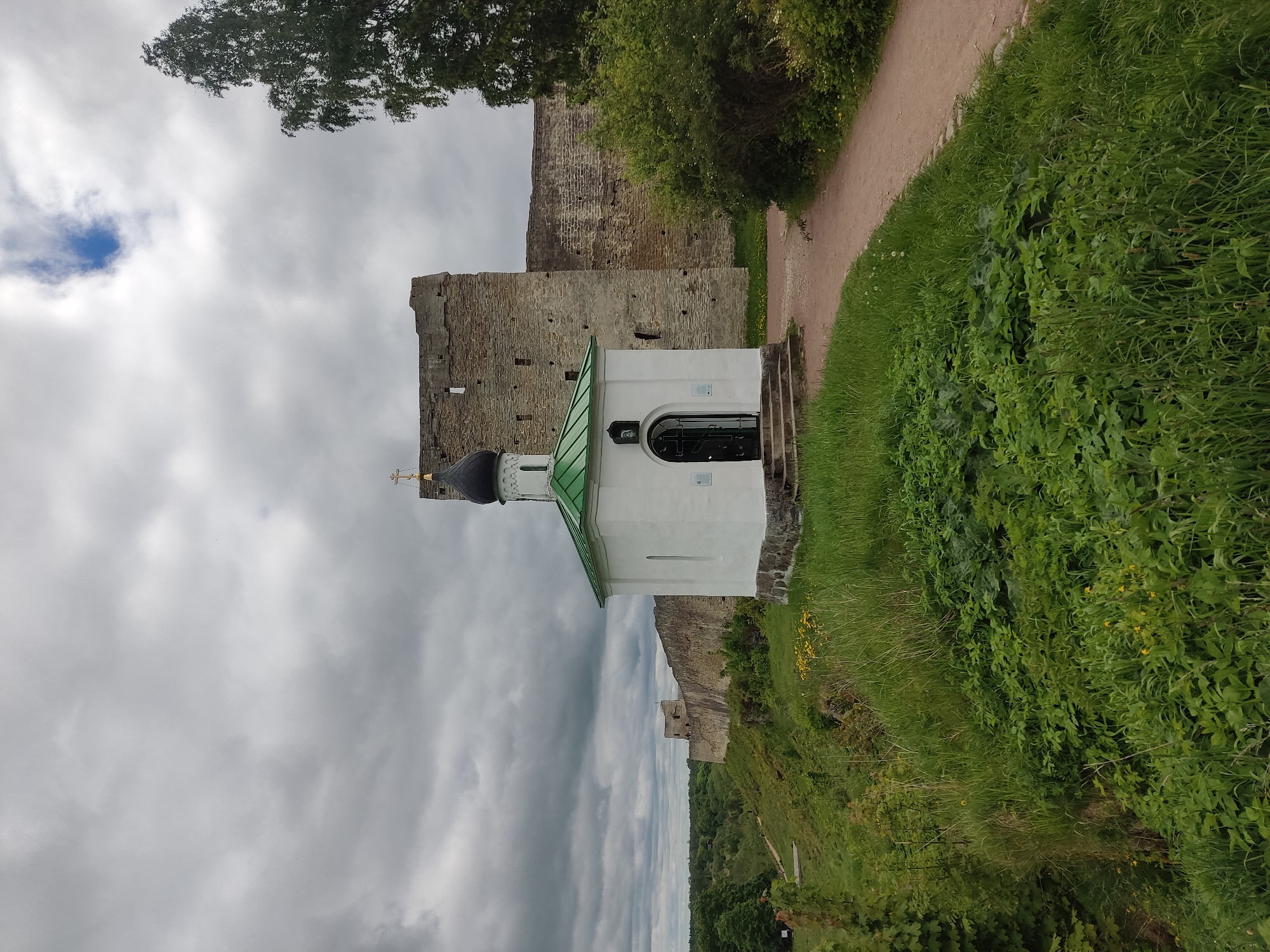 The image shows a small, white, single-story building with a green roof and a black onion dome topped with a golden cross. It's located beside a stone wall, presumably part of the Izborsk Fortress in Russia, due to the architectural style. A dirt path runs along a grassy area next to the wall. The sky is overcast with dark clouds. There are no people visible in the frame. Vegetation includes grass, leafy ground cover, and trees. The overall environment suggests a historical site or tourist attraction.