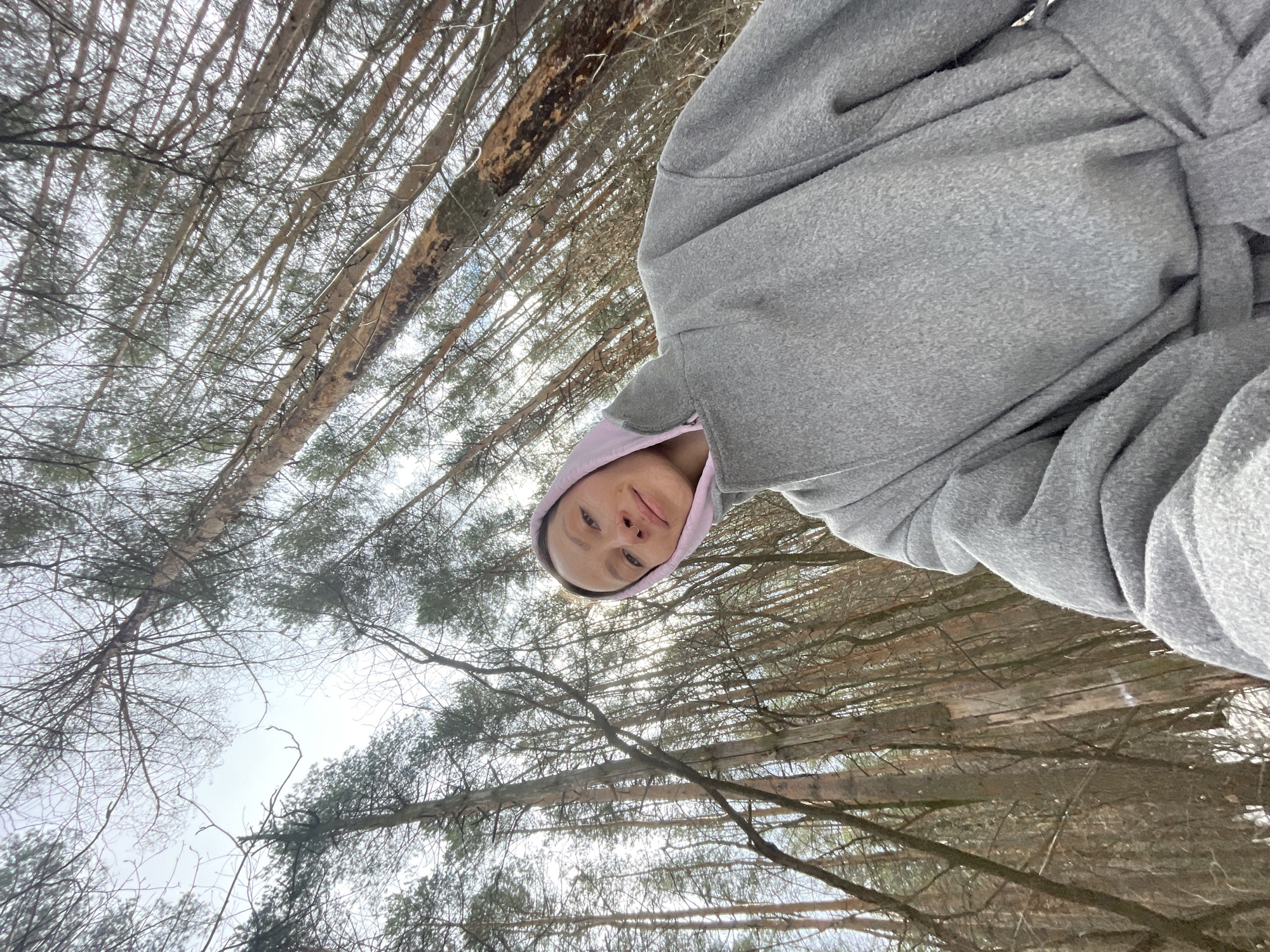 A woman wearing a gray robe and pink hoodie takes a selfie. She is outside in a forest of bare trees. The background is an overcast sky. It is not possible to determine if the location is in Izhevsk, Russia.