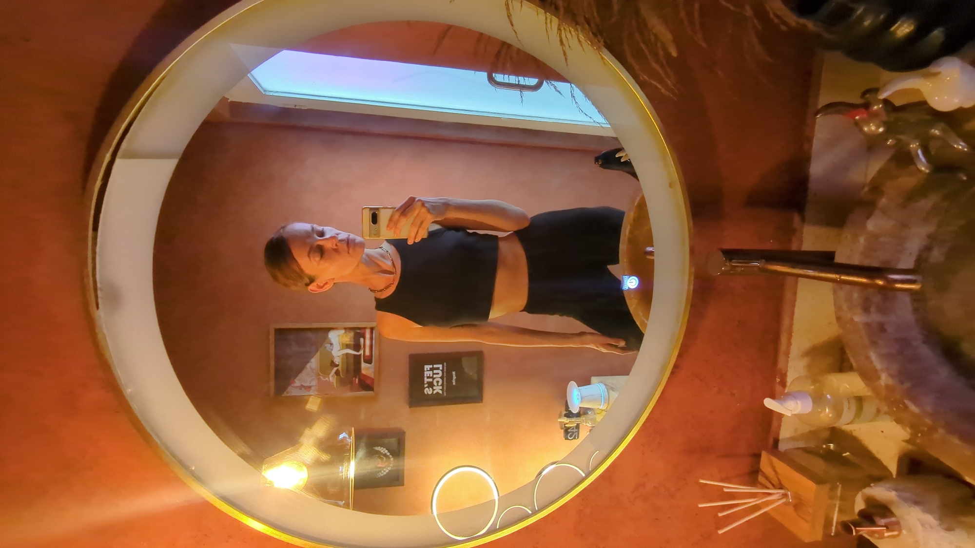 A woman in a black crop top and shorts is taking a selfie in a mirror. The mirror is round with a gold trim and a light around it. The mirror is located in a bathroom with orange-colored walls and decorations, including artwork and a soap dispenser. The bathroom is in North Kuta, Indonesia.