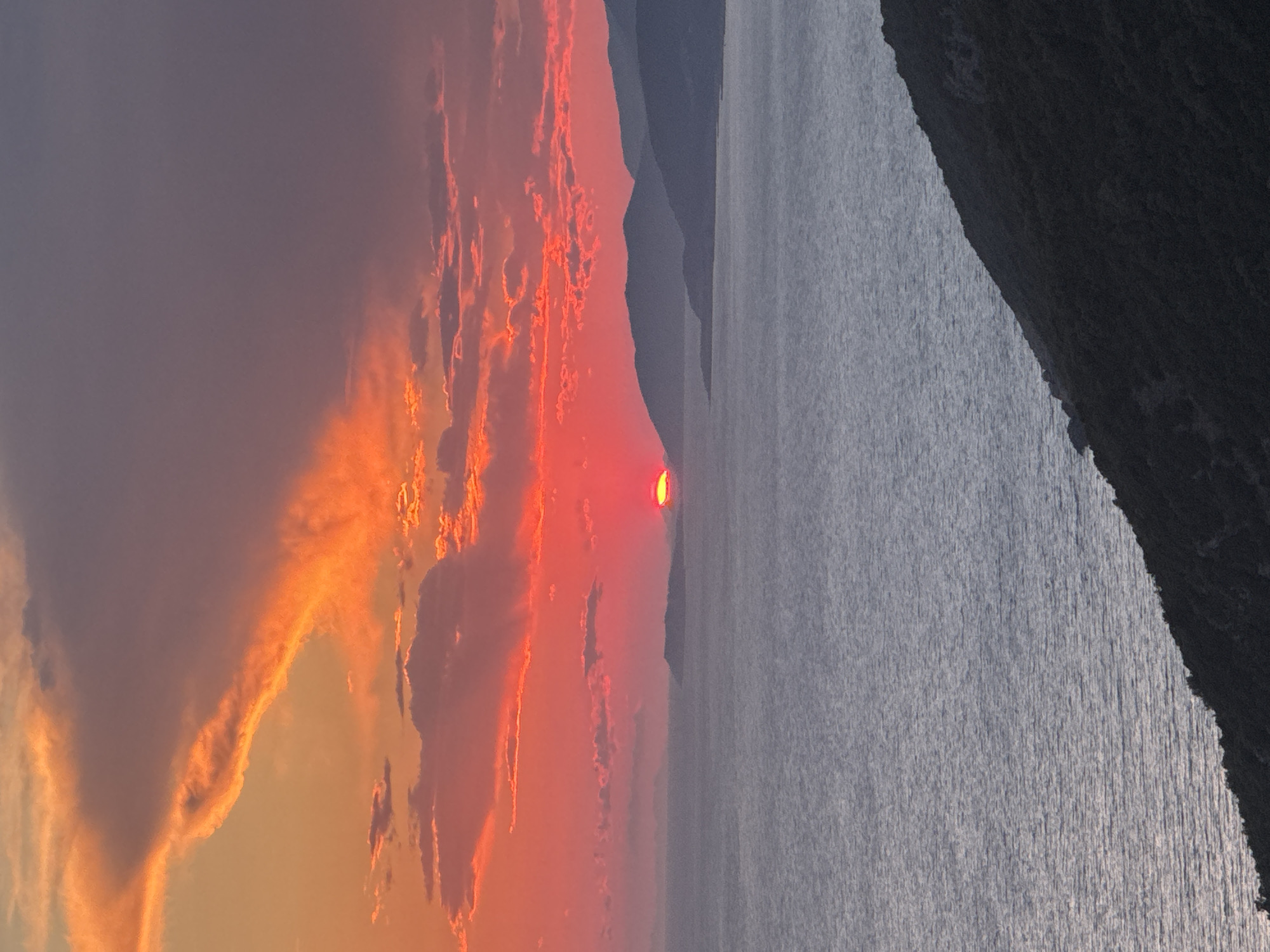 A scenic sunset is captured over a body of water, likely the Adriatic Sea, with mountainous terrain on the horizon. The sun, a vibrant red orb, is partially obscured by a distant island or landmass. The sky is ablaze with a dramatic display of orange, pink, and red hues, with wispy clouds reflecting the fiery colors. The water is calm, with subtle ripples, and the foreground is dominated by a dark, densely vegetated hillside. The scene evokes a sense of natural beauty and tranquility, typical of coastal Montenegro.