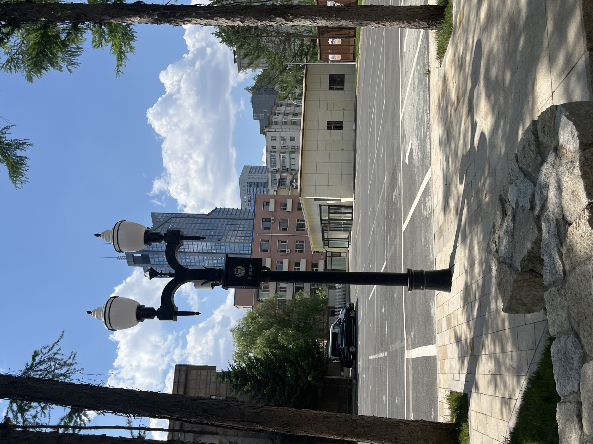 The image depicts an outdoor scene in Changchun City, China. A black street lamp with three white globe lights stands prominently in the foreground. The lamp has a sign on its pole with Chinese characters. Behind the lamp is a paved parking area with white painted lines, and several buildings of varying heights and architectural styles. A dark-colored sedan is parked along the edge of the parking area. Trees are visible, framing the top and left side of the view. A stone wall is on the right. The sky is blue with puffy white clouds.