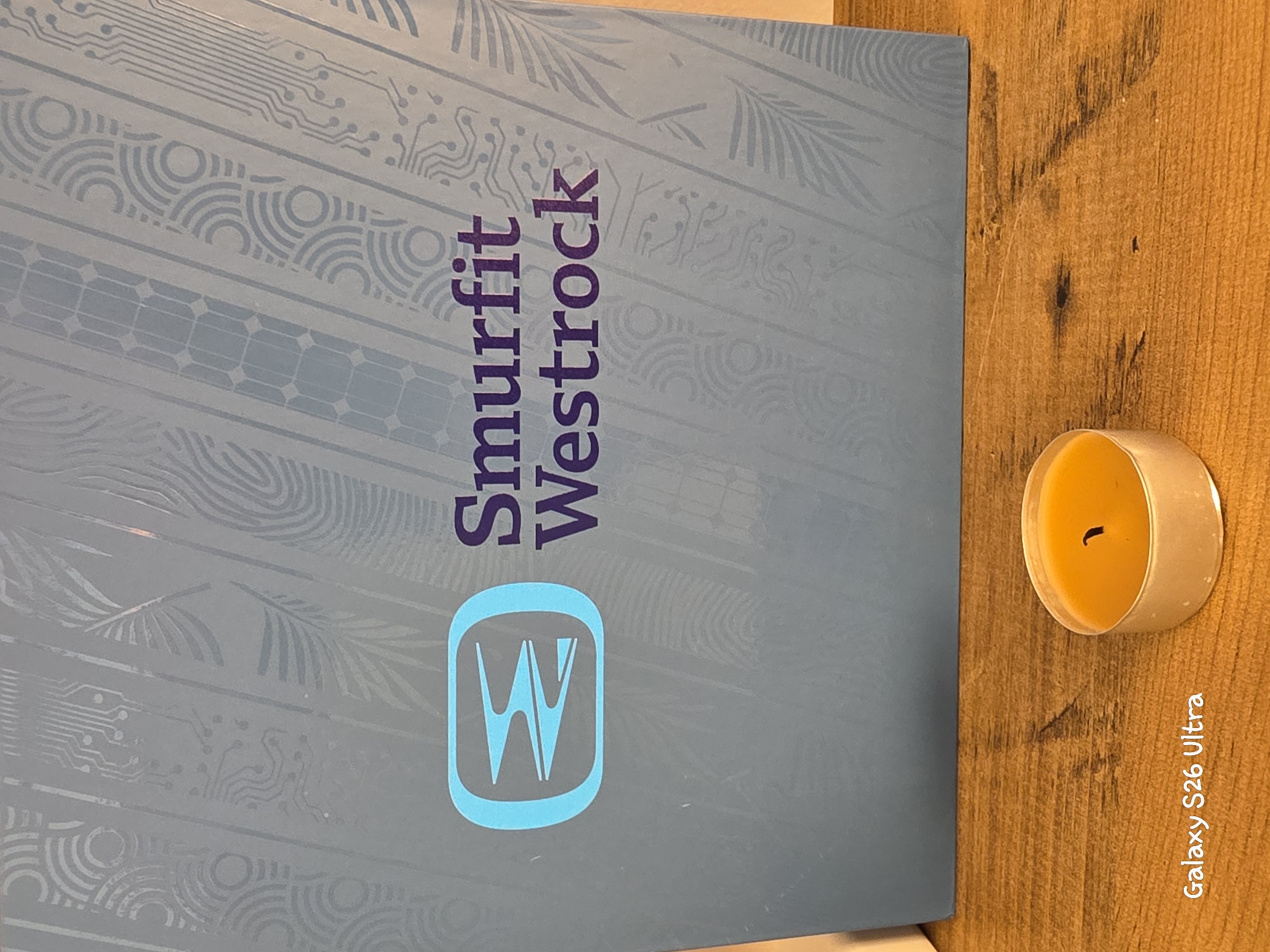 The image shows a close-up of a light blue box with a repeating pattern and the "Smurfit Westrock" logo in purple text. To the left of the text is a stylized, light blue logo. Below the box, on a wooden surface, is a small, lit tea candle in a silver holder. The bottom left corner of the image displays white text that reads "Galaxy S26 Ultra". The image does not provide any information about the city of Plovdiv, Bulgaria.
