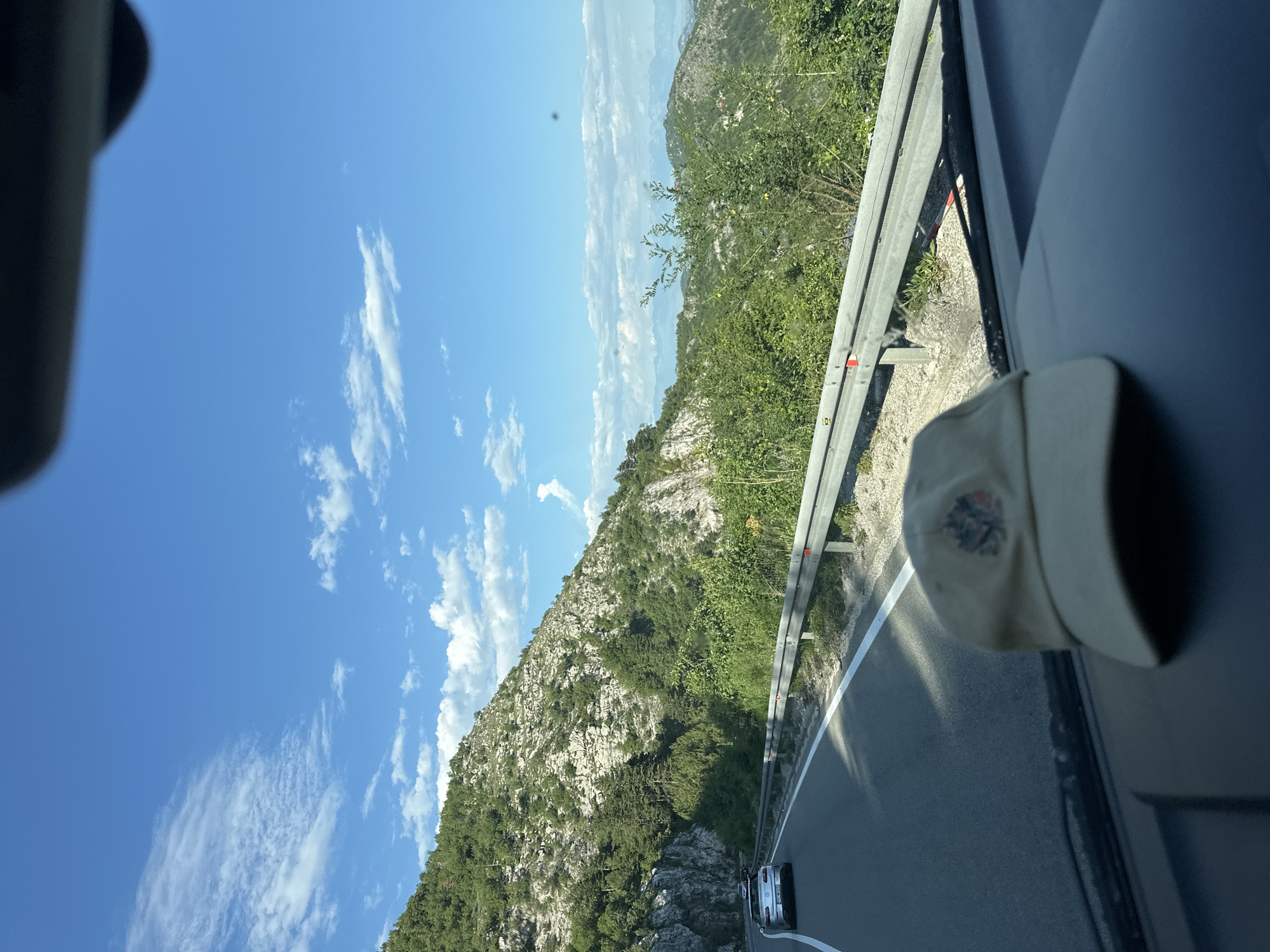The image depicts a view from inside a vehicle traveling on a road adjacent to a mountainside. The sky is blue with scattered clouds. The road is paved and has a guardrail. Vegetation covers the mountainside. A baseball cap is visible partially obscuring the view through the vehicle's window. A vehicle is visible in the distance on the road. Location is likely in or near Dobrsko Selo, Montenegro based on the landscape.