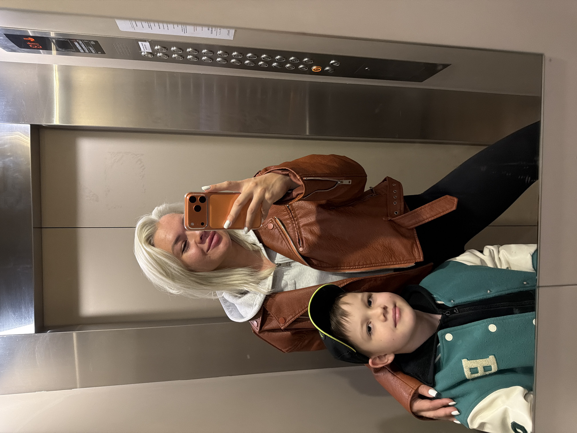 A woman and a young boy are taking a selfie in a mirrored elevator. The woman has blonde hair and is wearing a brown leather jacket over a grey hoodie. She is holding an orange mobile phone to take the picture. The young boy is wearing a green letterman jacket with a black cap. The elevator's control panel is visible in the background, showing floor numbers and buttons. The environment is enclosed and metallic.