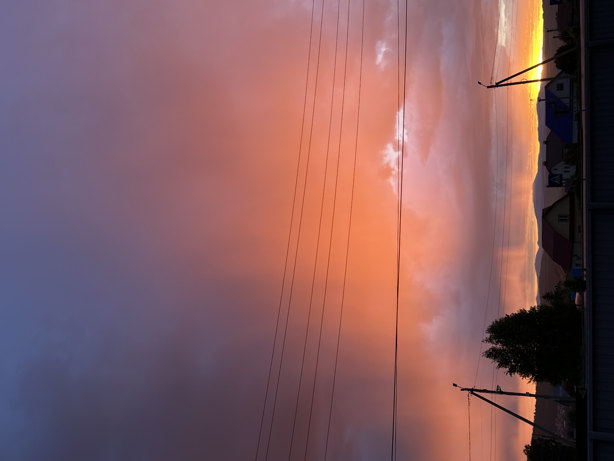 This media file captures a dramatic sunset or sunrise over a rural or semi-rural landscape. The sky dominates the upper two-thirds of the image, showcasing a vibrant gradient of colors. On the right, intense yellow and bright orange hues indicate the position of the sun, blending into warm pinks and coral tones across the mid-section. Towards the left, the sky transitions to cooler shades of deep purple and blue, suggesting the fading light. Fluffy white clouds are illuminated by the sun, appearing bright against the darker cloud formations.
Several black utility wires stretch vertically across the frame, cutting through the colorful sky. Two utility poles are visible towards the bottom of the image, holding these wires. The landscape below the sky is largely silhouetted against the bright light. Dark shapes of houses or buildings with varying rooflines, including one with a distinct blue roof, are visible along a distant horizon. Behind these structures, the silhouettes of mountains or large hills rise. A cluster of dark trees is also visible, and in the immediate foreground on the right, a dark fence or wall is present. The overall impression is one of natural beauty and a quiet, twilight scene. There is no visible text in the image.