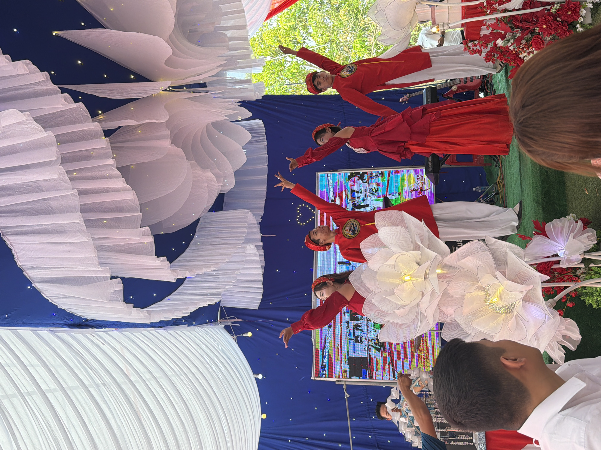This image captures a vibrant cultural performance taking place on a stage at an outdoor event, likely in Tay Son Commune, Vietnam. Four performers, dressed in traditional red Vietnamese attire, are the central subjects. Two men wear red áo dài-style tops with white pants, while two women wear red dresses or modified áo dài. All performers have their arms outstretched, indicating a dance or song routine, with one woman holding a microphone.
The stage is elaborately decorated. A deep blue backdrop, studded with small white lights resembling stars, forms the background. Large white ruffled fabric structures, reminiscent of clouds or abstract flowers, are dramatically draped on the left side of the stage. On the right, illuminated white flower-shaped lanterns add to the festive atmosphere. Behind the performers, a large screen displays colorful, abstract digital patterns and visuals, with the word "YAMAHA" faintly visible. Musical instruments, including a red bass guitar, are partially seen on the stage.
The scene is brightly lit by natural daylight, suggesting it is daytime. Green trees are visible in the background, confirming an outdoor or semi-outdoor setting. Red floral arrangements and white fabric draping further enhance the celebratory mood. In the foreground, the back of a person's head and shoulder are visible, looking towards the stage, indicating an audience member. The overall event appears to be a lively and engaging cultural celebration.