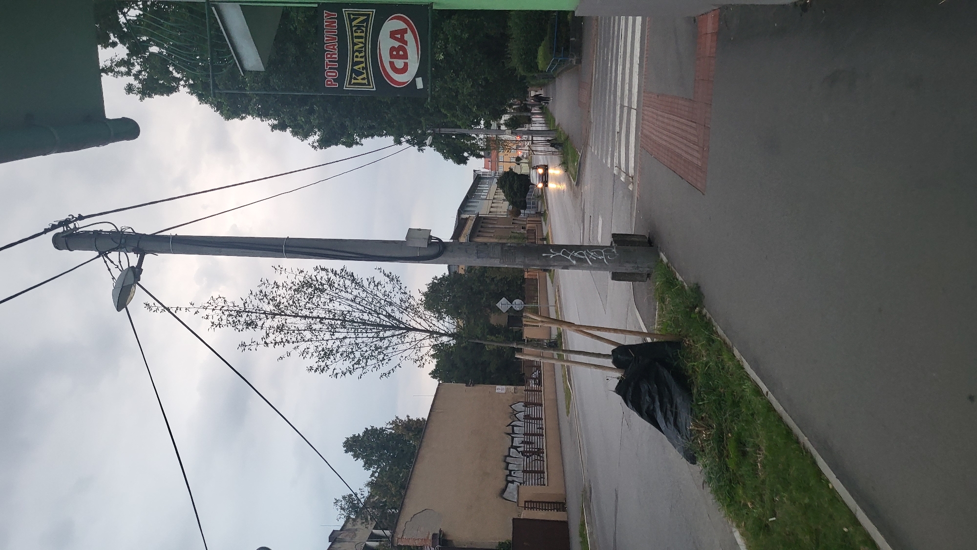 The image depicts a street scene in Prešov, Slovakia, likely on a cloudy day. In the foreground, a gray sidewalk runs parallel to a street. To the left, a small tree is covered in black plastic, supported by wooden stakes. A street light pole with electrical wires is prominent in the mid-ground. Across the street, buildings are visible, including one with graffiti on its fence. A crosswalk is present on the right side of the image, leading to a sidewalk. A car with its headlights on is driving down the street in the distance. A sign for "POTRAVINY KARMEN CBA" is mounted on a building on the right. A person is walking on the sidewalk on the right. The overall environment suggests an urban setting with elements of infrastructure and commerce.