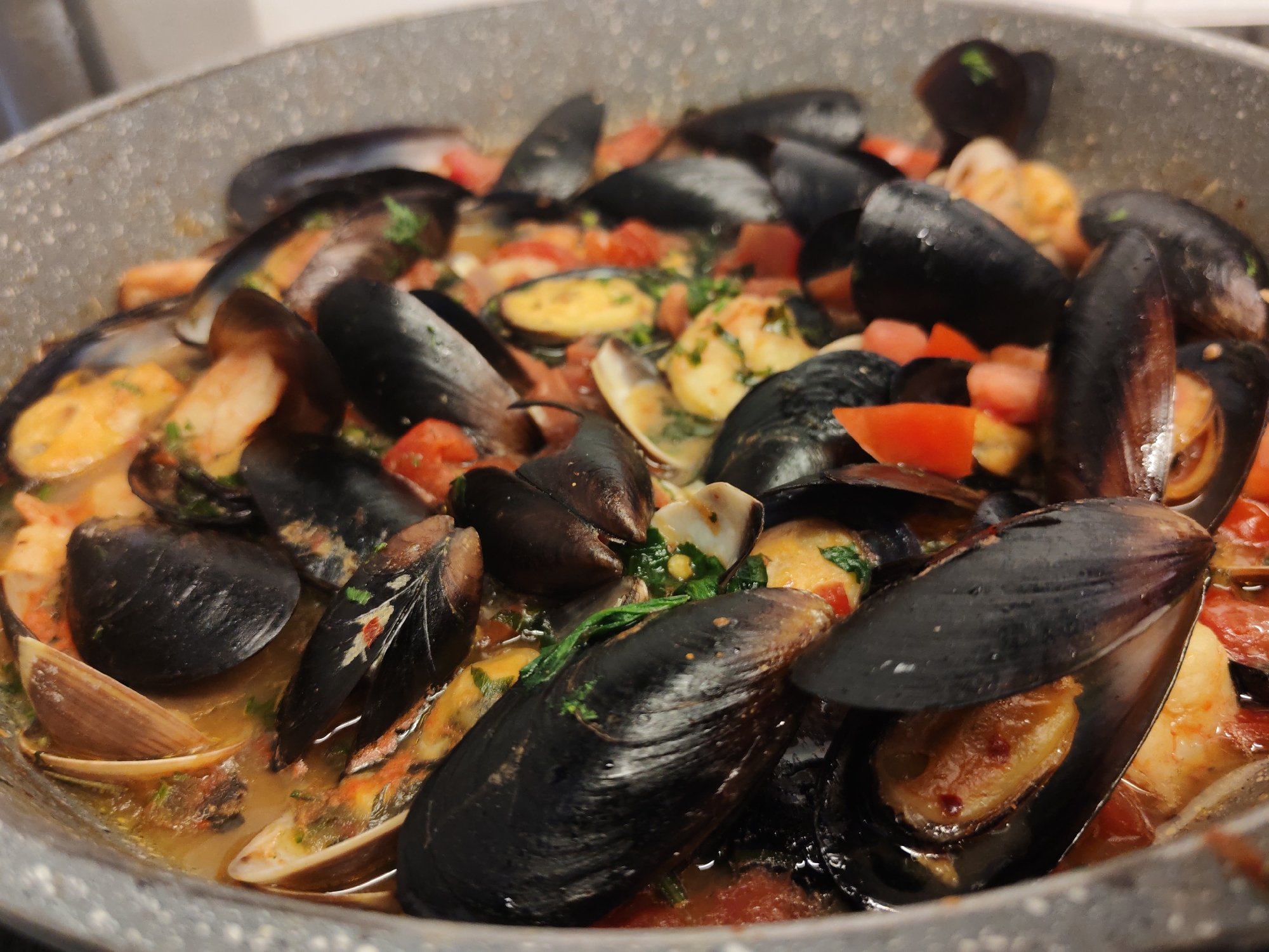 The image shows a close-up, overhead view of a pan filled with cooked mussels and other seafood in a sauce. The mussels are dark blue-black and many are open, revealing the orange-yellow flesh inside. Chunks of red tomato and green herbs are visible amongst the mussels. There are also some pinkish shrimp scattered throughout the dish. The pan appears to be made of a dark gray material with a slightly textured surface. The background is out of focus but suggests a kitchen environment. The dish is likely a seafood stew or marinara, a common preparation in coastal Italian cuisine. While this image depicts a dish commonly enjoyed in La Spezia, there is no specific information or visual cues to confirm its location within the city.
