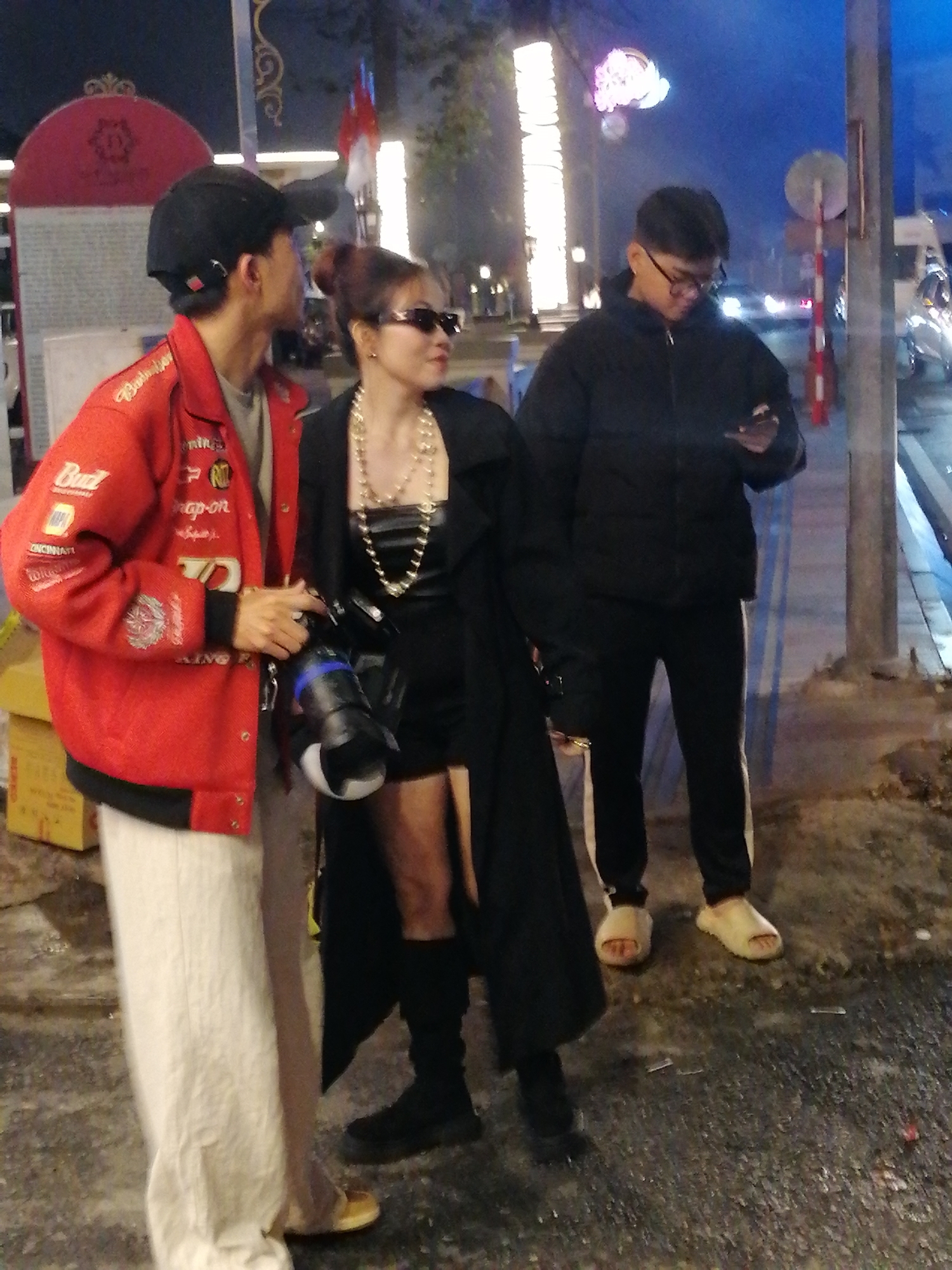 The image captures a street scene in Da Lat, Vietnam at night. Three individuals are visible. On the left, a person wearing a red jacket with various patches and a black baseball cap is facing away from the camera, looking to their left. They are wearing loose, light-colored pants. In the center, a woman with dark sunglasses and a black coat over a black outfit stands facing the camera, holding a large camera with a lens in her right hand. A multi-layered pearl necklace is draped around her neck. To the right, a young man wearing a black puffer jacket and black pants with white stripes is looking down at his phone. He is wearing glasses and light-colored slides on his feet. The background suggests a city street with artificial lighting, including illuminated buildings and streetlights, and indications of traffic.
