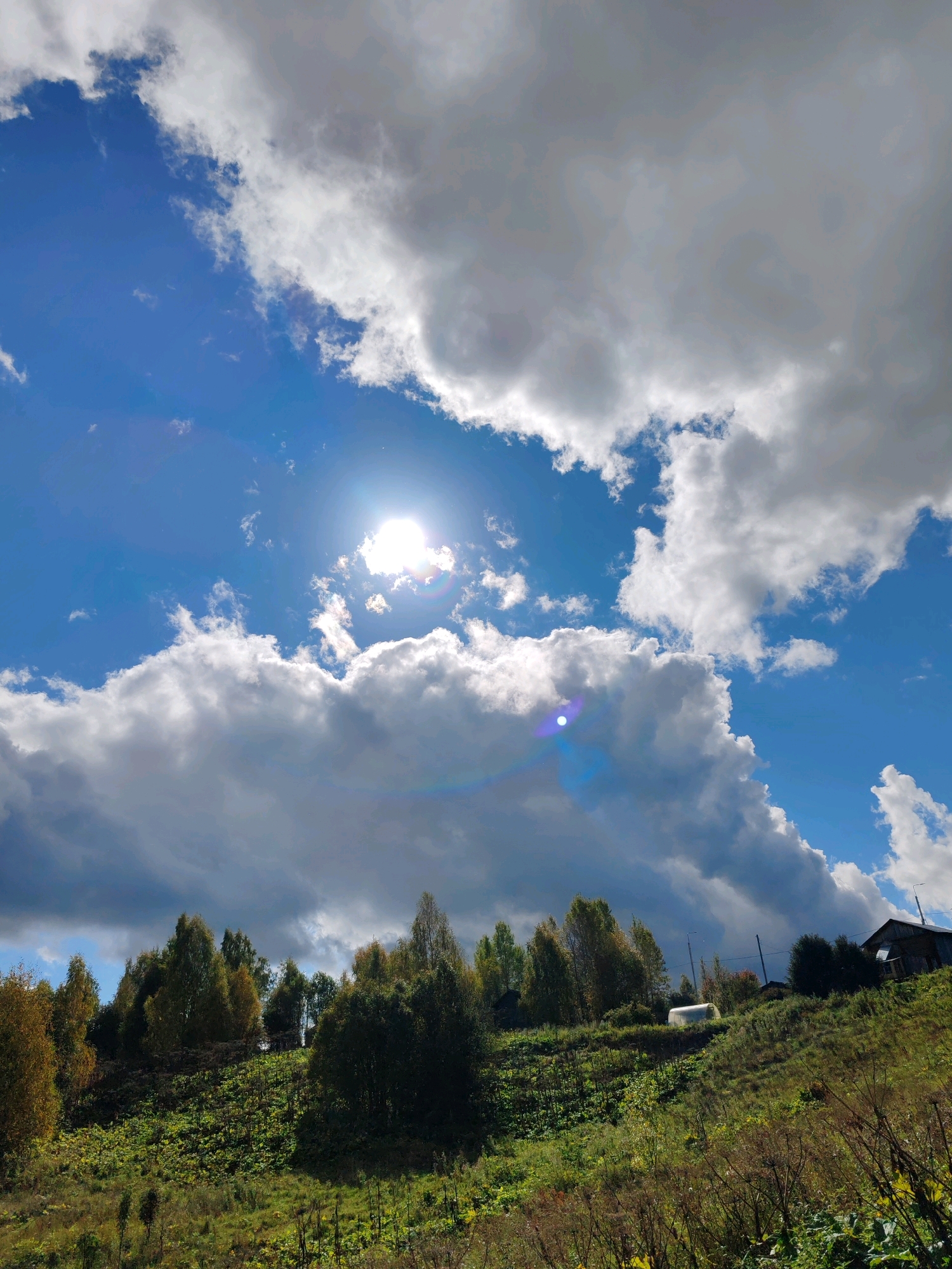 The image depicts a rural landscape with a bright, sunlit sky filled with fluffy white and gray clouds. The sun is visible, casting a lens flare. In the foreground, a grassy slope is covered with lush green vegetation and some dry plants. A line of trees with autumnal foliage is situated on the crest of the hill. On the right side of the hill, a wooden building with a dark roof and a smaller white structure, possibly a greenhouse or shed, are visible. The scene suggests a natural environment in a rural or suburban area, likely in Чухлэм, Russia, given the context. There are no people visible in the image.