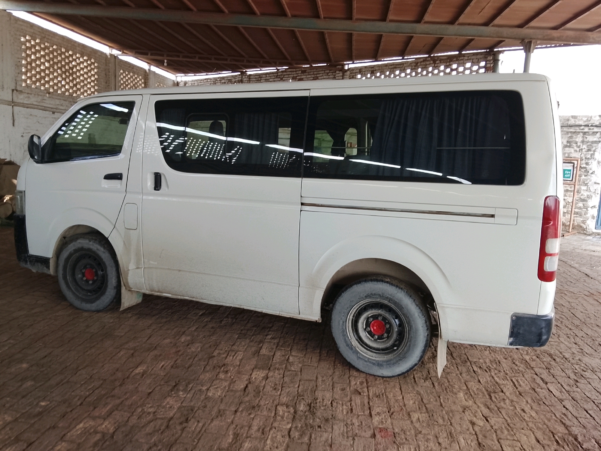 A white van is parked under a covered structure in Pataar Goth, Pakistan. The van is facing towards the right of the image and has tinted windows with dark curtains visible inside. The exterior of the van appears dusty. The ground beneath the van is paved with dark brown bricks. The structure above the van has a wooden roof with visible beams. To the left of the van, a white brick wall with patterned openings is visible, allowing light to filter through. A sign with "fire exit" in English is partially visible on a wall in the background to the right.