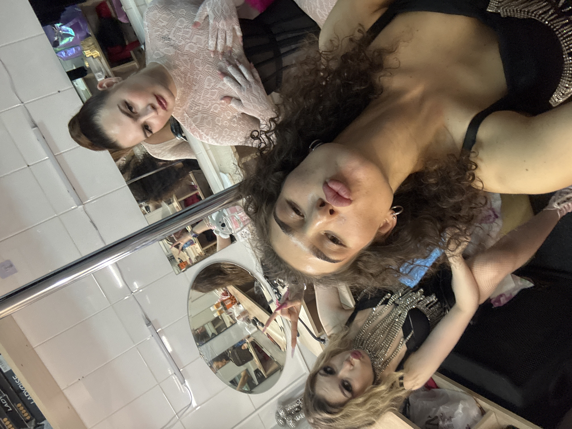 Three women are posing for a selfie in a dressing room. The woman in the foreground has curly brown hair and is wearing a black top. She is making a duck face. To her left, a woman with blonde hair is also making a duck face and wearing a black top with a large, jeweled necklace. Behind them, a woman with her hair in a bun is wearing a white, lace-like top. The room appears to be a backstage dressing area, with mirrors, shelves, and lighting visible. There are items such as makeup and costumes on the shelves. The image is taken from a close-up perspective, focusing on the women's faces and upper bodies.