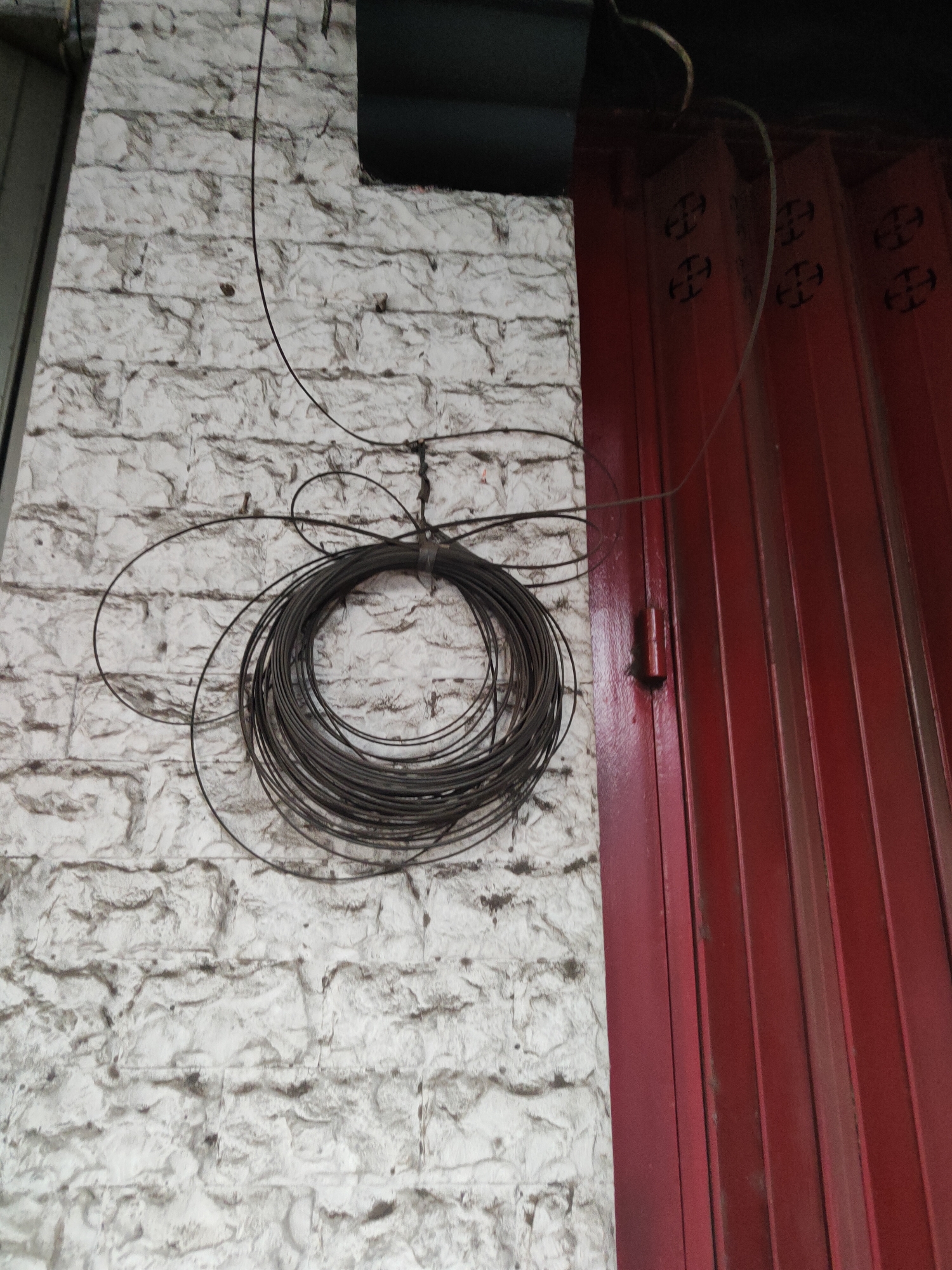 The image depicts an outdoor scene in Bangil, Indonesia, featuring a textured white brick wall on the left and a red corrugated metal gate on the right. A tangled coil of dark wire is suspended from the wall by what appears to be a tie. The wire is thick and appears to be coiled into a circular bundle, with loose ends trailing away. The brick wall has a rough, uneven surface with visible mortar lines. The red gate shows vertical pleats or corrugated lines and has a slightly worn appearance. The overall lighting suggests it is daytime. There are no people visible in the image.