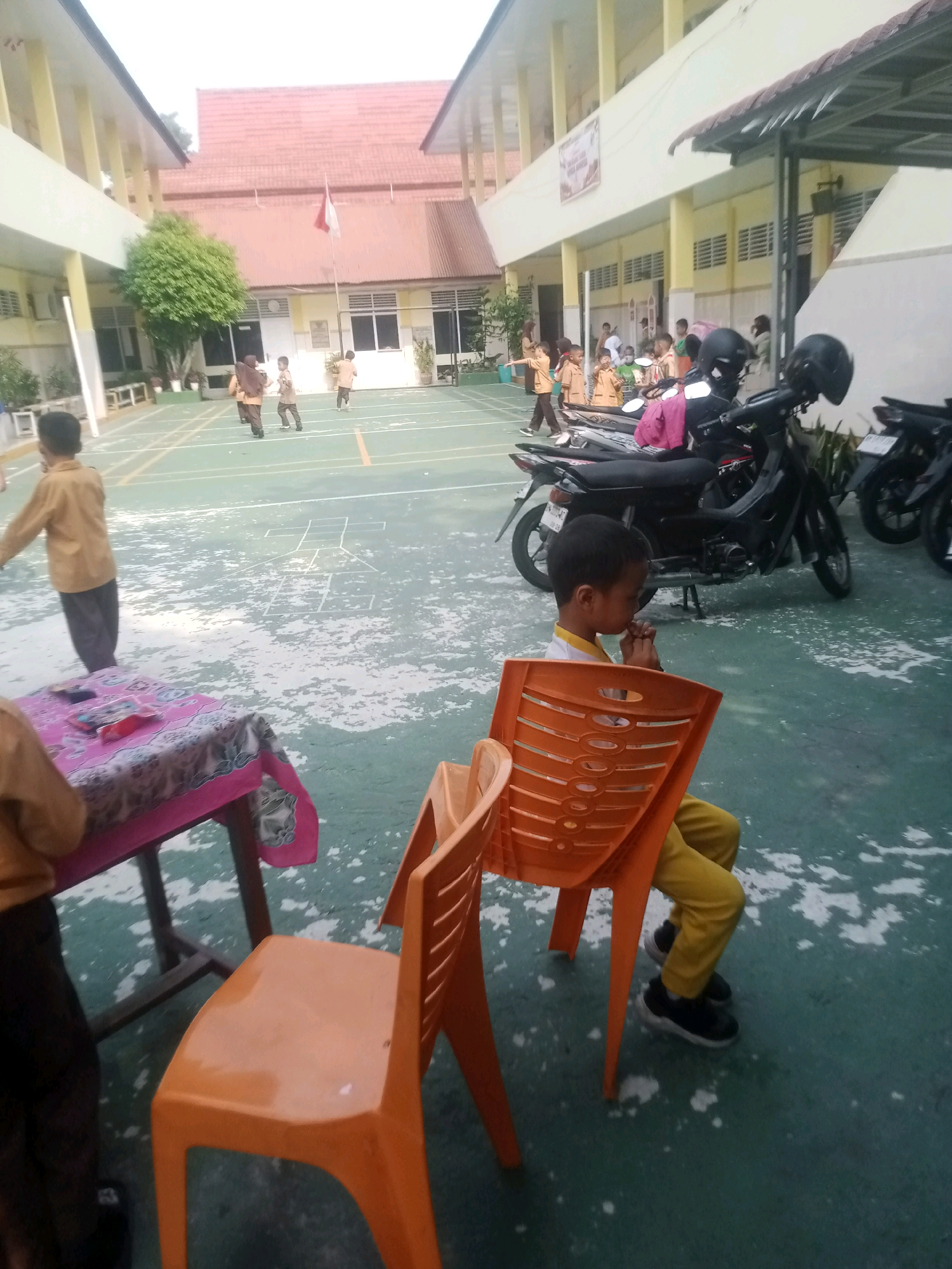 A group of children are playing on an outdoor basketball court at what appears to be a school. The court is green with white lines, and a hopscotch grid is drawn on the ground. In the background, a building with a red tile roof and yellow exterior walls is visible. There are several motorcycles parked near a covered walkway. One child in the foreground is sitting on an orange plastic chair, looking down with their hands clasped. Another child, wearing a tan uniform, is walking away from the camera. The scene is likely from a school in Pekanbaru, Indonesia, given the architectural style and the presence of motorcycles.