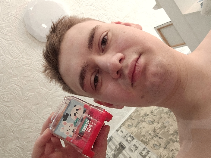 A fair-skinned person, likely male, with short brown hair, holds a small, red-framed container with a cartoon animal design. The container appears to hold markers or pens. The background features a textured white wall with a rounded light fixture, and a glimpse of a ceiling and wall corner suggesting an interior setting. The photo is taken at a close, angled perspective.