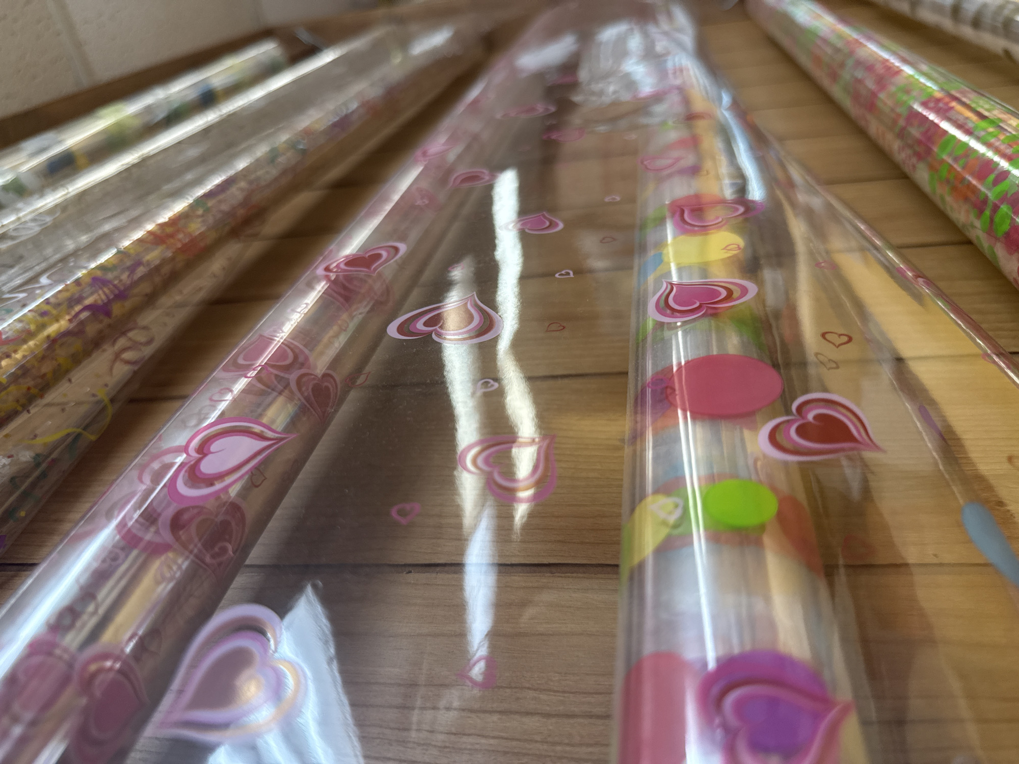The image shows a close-up view of several rolls of wrapping paper with different patterns. The rolls are arranged horizontally on what appears to be a wooden shelf or surface. The focal point is on the middle roll, which features a clear plastic wrapping with pink heart designs. Other rolls with varied colors and patterns are partially visible on either side. The scene is indoors, likely in a store or storage area. The location is Davie, United States of America.