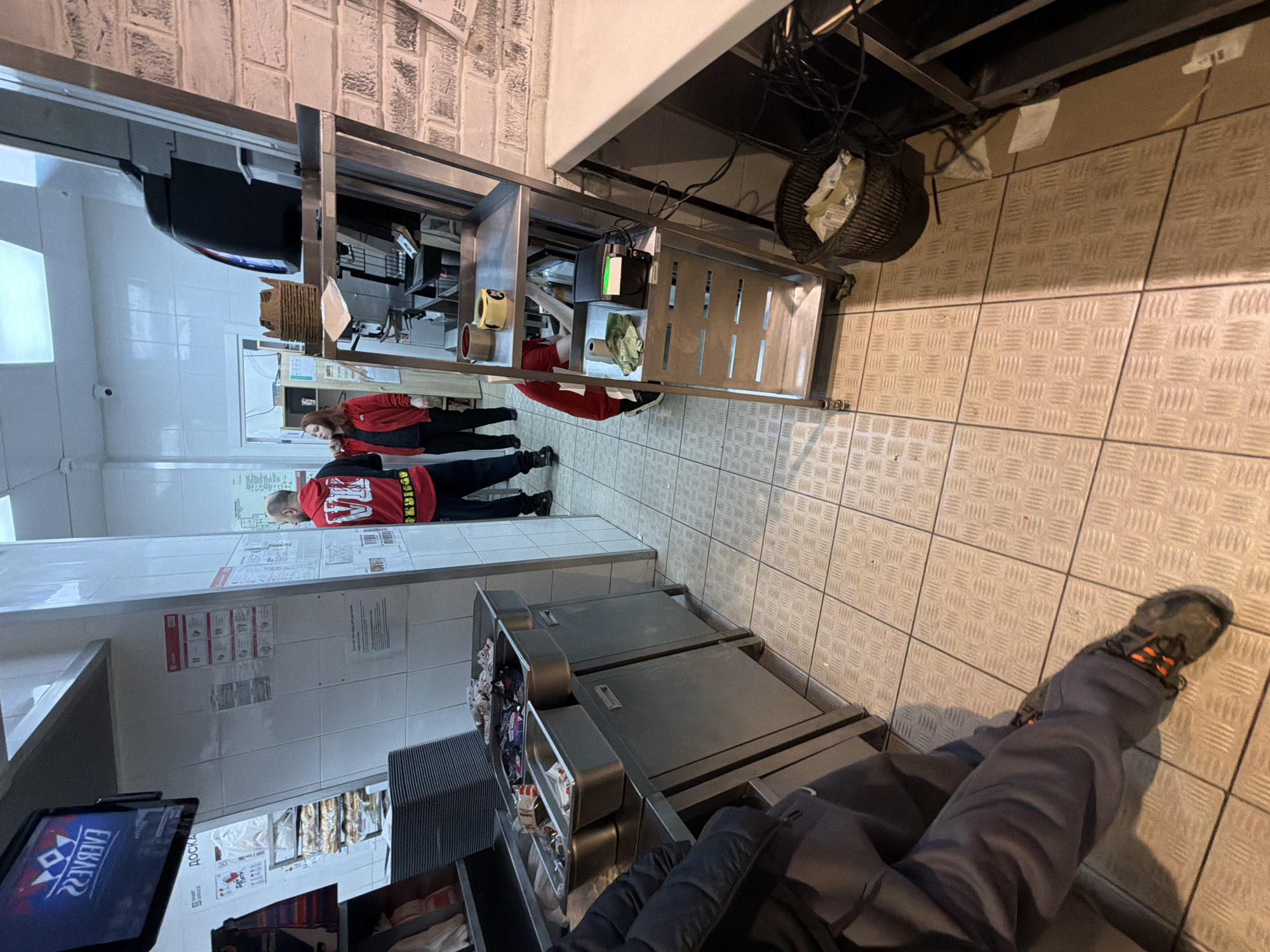 A high-angle, rotated view of what appears to be a fast-food restaurant kitchen, potentially in Chaikovsky, Russia. Two individuals are visible in the background wearing red shirts with black pants. One person seems to be working or leaning over a stainless steel counter, while the other individual is standing upright. The foreground shows the legs and feet of a person wearing gray pants and hiking shoes, who is likely the photographer. Nearby are stacked, stainless steel food preparation stations holding various food items. A screen displaying branding is visible lower left. The floor is covered in diamond-patterned gray tiles, and overhead there is a brick wall. Various wires and a waste basket are visible beneath an overhanging structure.