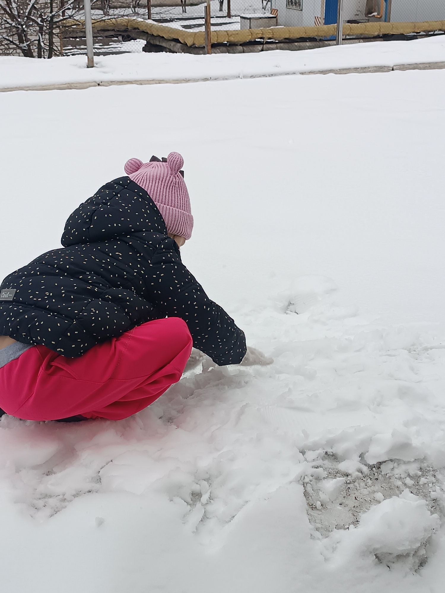A child in a pink hat and red pants is crouched in the snow. The child is wearing a dark jacket with white specks. The scene is set outdoors with snow covering the ground. In the background, there are trees and a building. The city is Unknown, Unknown.