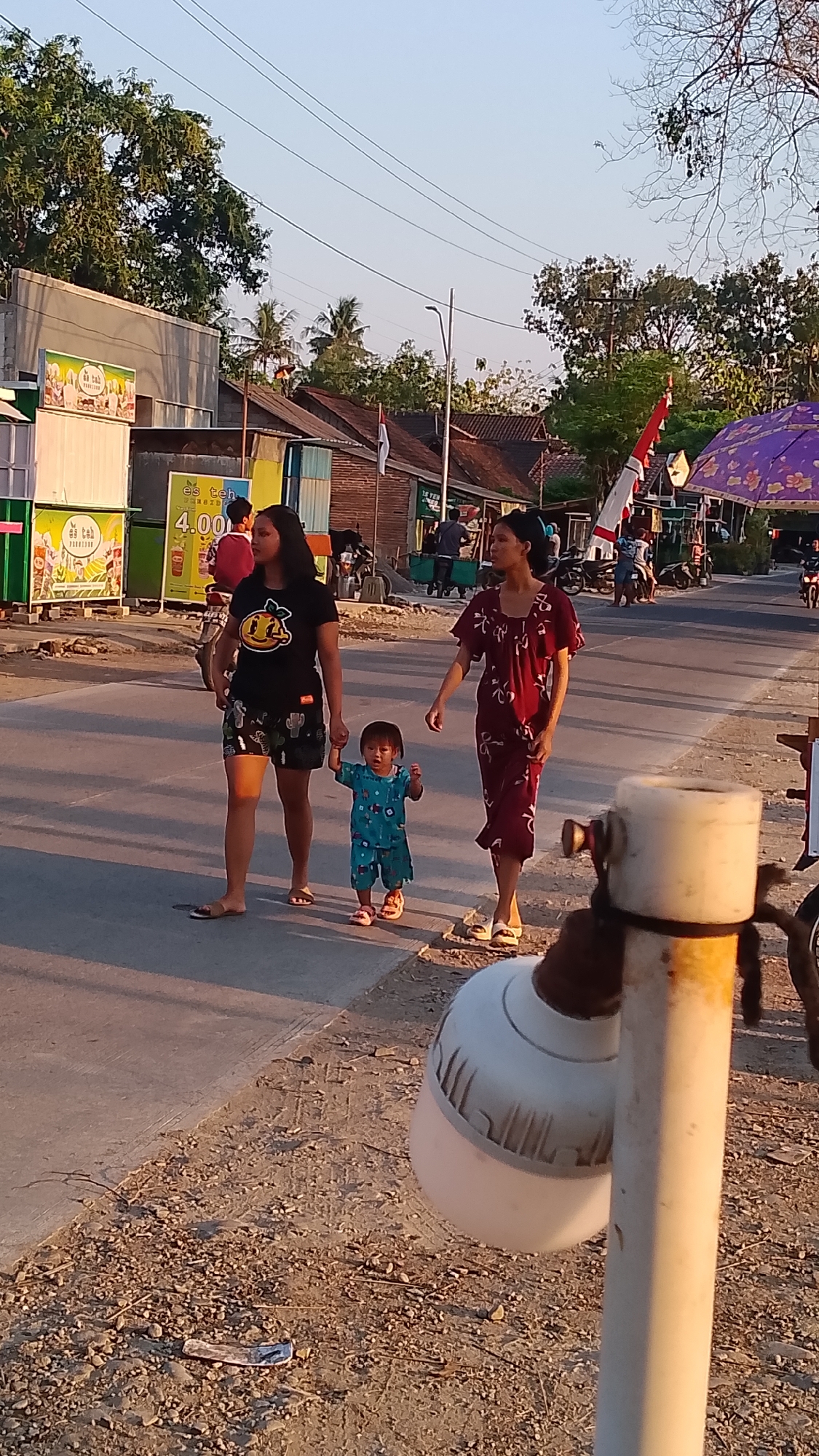 In Karanglangu, Indonesia, two women and a young child walk along a paved road during the day. The woman on the left wears a black t-shirt with a cartoon face and patterned shorts, while the woman on the right is in a floral patterned dress. The child, dressed in a blue outfit, walks between them, holding the left woman's hand. The road is lined with shops and trees, and power lines are visible overhead. A purple umbrella is set up on the right side of the road, and several motorcycles are parked nearby. The ground is a mix of dirt and gravel. A white lamp fixture is prominently displayed in the foreground.