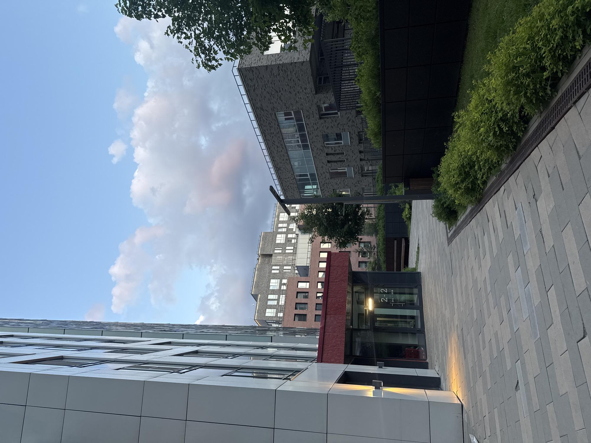 This image captures a modern architectural complex in Khimki, Russia, featuring multiple buildings, a paved walkway, and landscaping under a partially cloudy sky. The overall scene suggests a contemporary urban or suburban environment, possibly a residential or commercial development.
In the foreground on the left, a tall, modern building with a light grey or white panel facade and numerous large, rectangular windows reflects the sky. Adjacent to it, illuminating the paved path, are warm-toned artificial light fixtures. Further along the pathway, a glass entrance is visible, displaying the text "2-2" and "251-356".
To the right and slightly further back, a darker brick building stands, featuring various window sizes and a section with reddish-brown panels. Beyond this, another multi-story building with a facade of varied shades of grey and brick is visible in the background. Towards the upper right, a dark-faced building with lush green bushes and trees along its base and a green roof is present, creating a contrast with the urban structures.
The ground is covered with a wide, grey paved walkway made of rectangular stones, which leads into the distance. A metal drainage grate runs along the edge of this path. Green bushes and hedges line the walkway and the bases of some buildings, adding natural elements to the scene. Overhead, the sky is a clear blue with scattered white and grey clouds, some exhibiting a subtle pinkish tint, suggesting it is either late afternoon or early evening. The lighting is soft and natural, with shadows indicating a low sun angle. No people or specific activities are visible, presenting a serene and unoccupied environment.