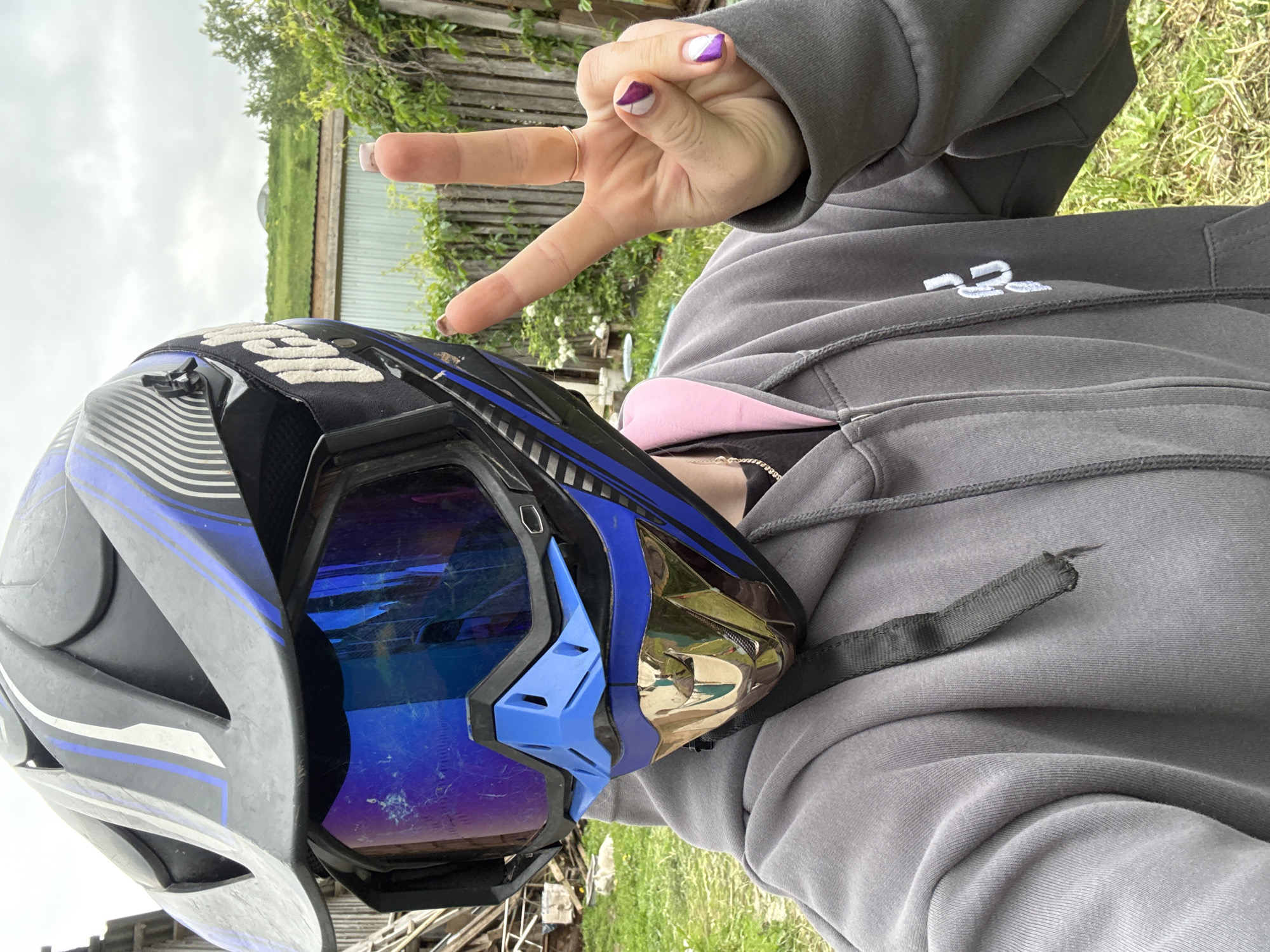 An individual is depicted outdoors in a semi-rural setting within the vicinity of Ляльшур, Russia, from a first-person perspective. The person is wearing a gray zip-up hoodie with a pink lining visible in the hood, and a dark strap hangs from their left shoulder. A subtle white logo is present on the right chest area of the hoodie. A gold chain necklace is visible around the neck. The subject's head is fully obscured by a black, blue, and gray motocross-style helmet featuring a large visor and a blue and gold/bronze chin guard. The helmet is adorned with blue and white linear patterns. Blue mirrored goggles, exhibiting visible scuffs and scratches, cover the eyes. The goggle strap, partially visible, displays white text. The person's right hand is raised, forming a peace sign with the index and middle fingers extended. A plain thin gold ring is worn on the right ring finger. The fingernails are manicured: the thumb, index, and middle fingers are painted a solid dark brown/mauve color, while the ring finger and pinky feature a diagonal split design, with one half being white and the other a metallic purple/pink. The background features green grass and sparse vegetation in the immediate foreground, transitioning to a weathered wooden fence. Beyond the fence, an unkempt grassy area with various bushes and a dilapidated structure with a corrugated metal roof is visible. In the distant background, under an overcast sky, a green hill is prominent, topped by a subtle, light-colored spherical or domed structure.