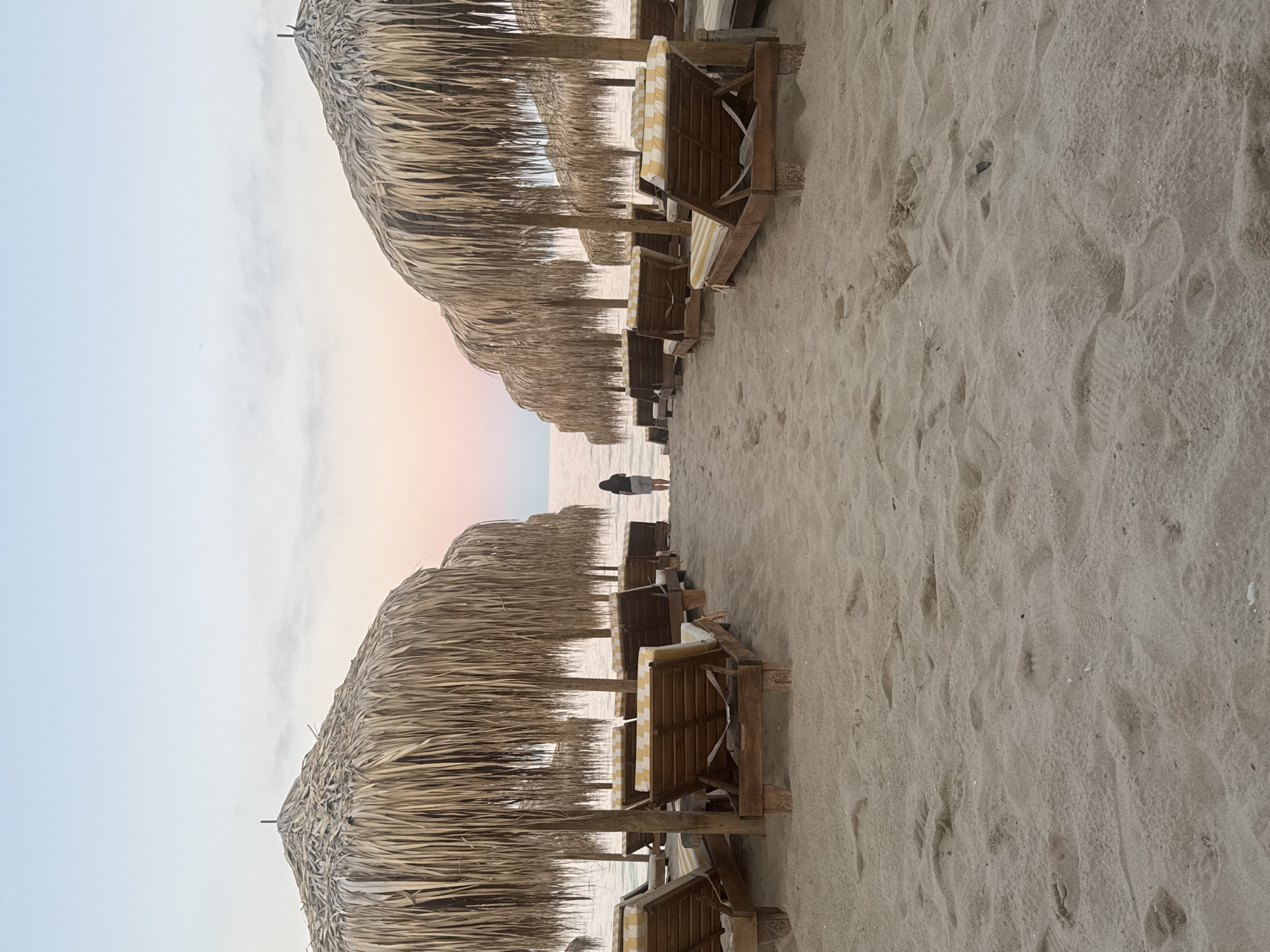 In Kranevo, Bulgaria, on a sandy beach at dusk, two rows of empty, sun-bleached thatched umbrellas face the sea. Beneath the umbrellas are wooden lounge chairs with yellow and white striped cushions. A lone figure, seen from the back, stands at the water's edge between the rows of umbrellas, facing the calm sea and the pastel-colored sky. The sand in the foreground is imprinted with footprints.