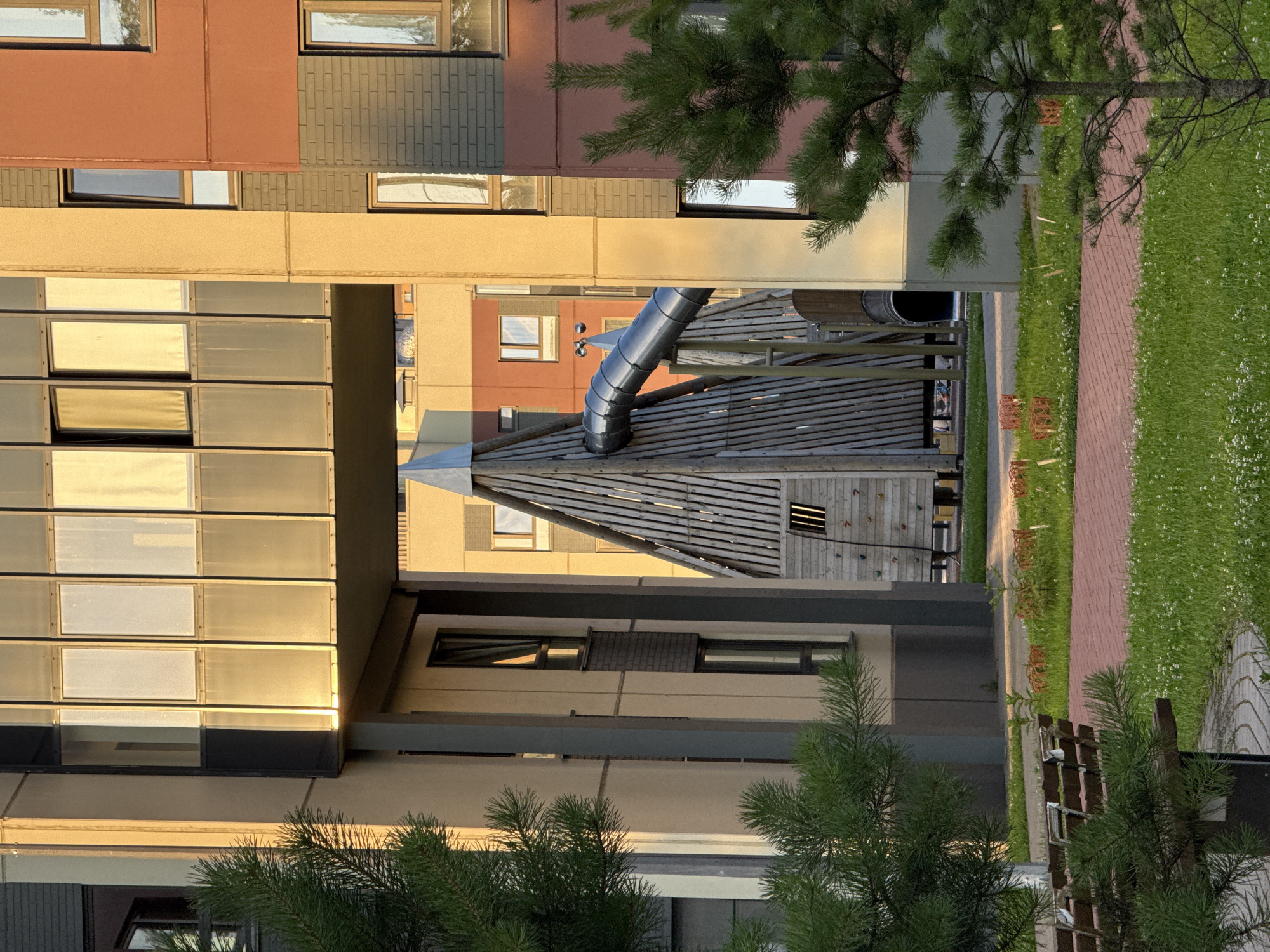 In Pargolovo, Russia, a modern residential complex features an elevated walkway connecting two buildings, casting a shadow over a children's play structure. The structure is made of wood with a metal slide and a climbing wall. Pine trees frame the scene, and a well-maintained courtyard with grass, brick paths, and benches suggests a park-like setting. The warm evening sun bathes the buildings in a golden light, highlighting the contemporary architecture.