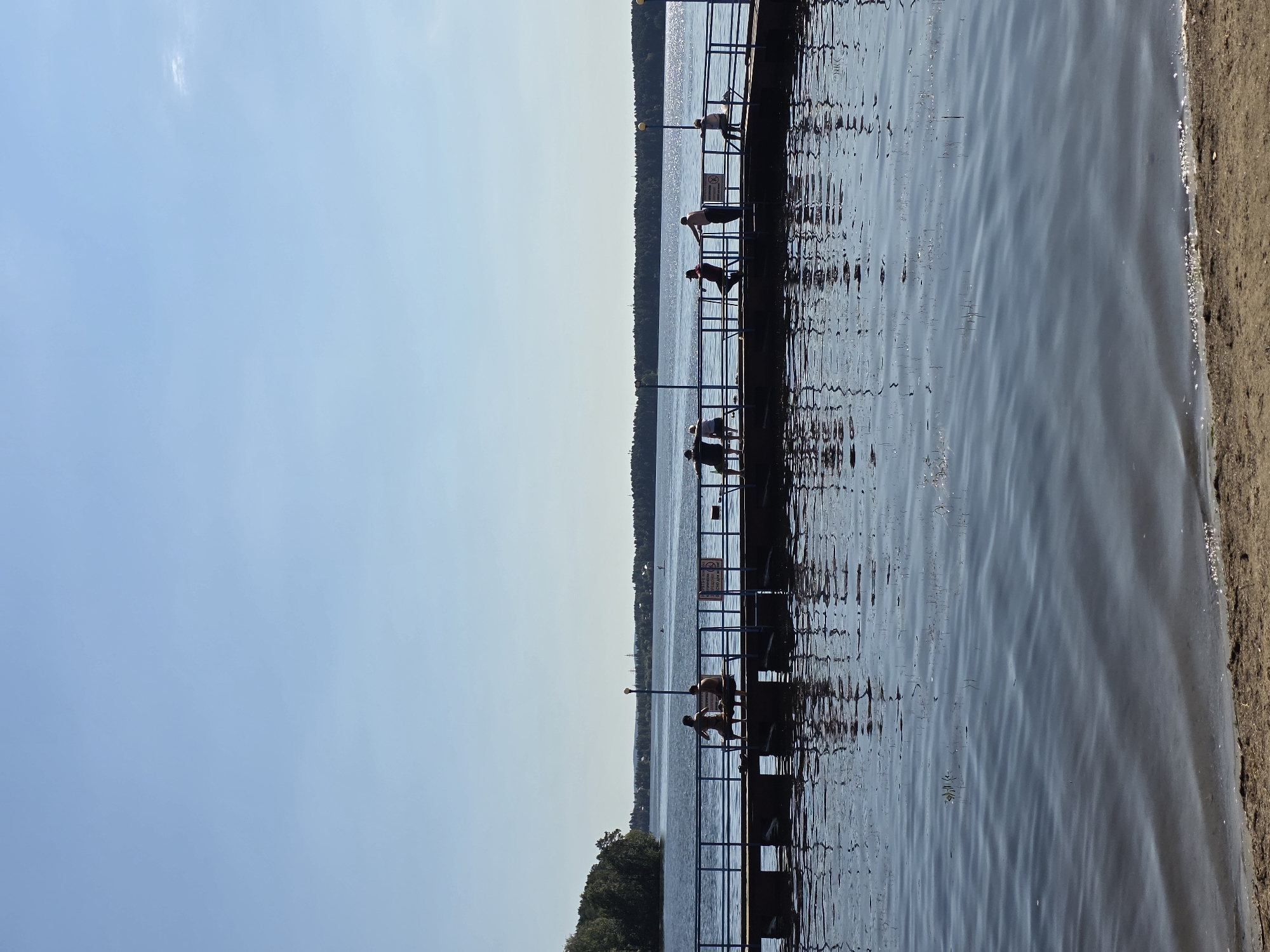 The image depicts a sunny day at a pier on Lake Chebarkul, located in the city of Chebarkul, Russia. A wooden pier extends into the calm, blue water, with a blue railing running along its length. Several people are visible on the pier, some sitting and others standing, enjoying the view or relaxing. Silhouettes of people are also reflected in the water. The background features a tree-lined shore and a clear blue sky. The foreground shows a sandy beach and the gentle ripples of the lake.