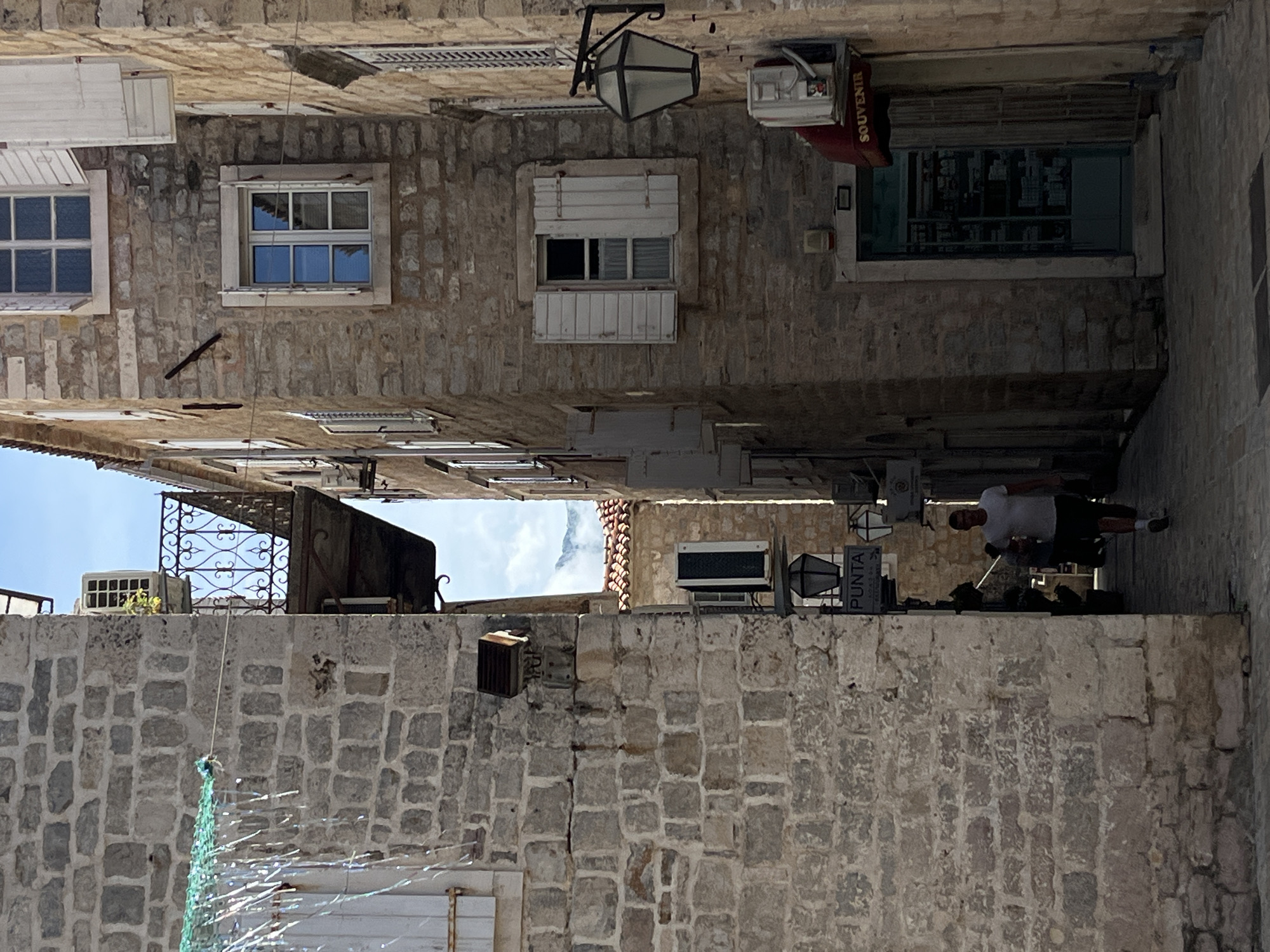 The image depicts a narrow, sunlit alleyway winding between tall, aged stone buildings, characteristic of a historic town. The architectural style features rough-hewn stone blocks for the walls, giving the scene a rustic, ancient feel. A clear blue sky is visible in the strip above the buildings, indicating bright daylight.
On the left side, the building displays a mix of windows; one near the top has closed white louvered shutters, while below it, another window with white frames is open, revealing multiple glass panes reflecting the sky. Further down, at street level, a stone wall is visible, partially obscured by a bright green, netted object and shimmering light, possibly from a small fountain or reflecting water. An air conditioning unit and an ornate metal railing are perched on a rooftop in the upper left background.
The building on the right side of the alley also features windows with white shutters, some closed and one partially open. A classic black lantern, with a hexagonal glass design, hangs from the wall, adding to the old-world charm. Below this, a small shop is indicated by a red awning bearing the white text "SOUVENIR," suggesting a tourist-oriented business. Further into the alley, a white sign with black lettering prominently displays "PUNTA." A single person is walking away from the viewer in the distance, dressed in a light-colored t-shirt, dark shorts, and sneakers, their figure providing a sense of scale to the passage. The time of day appears to be bright daytime, with clear skies and good illumination throughout the alley.