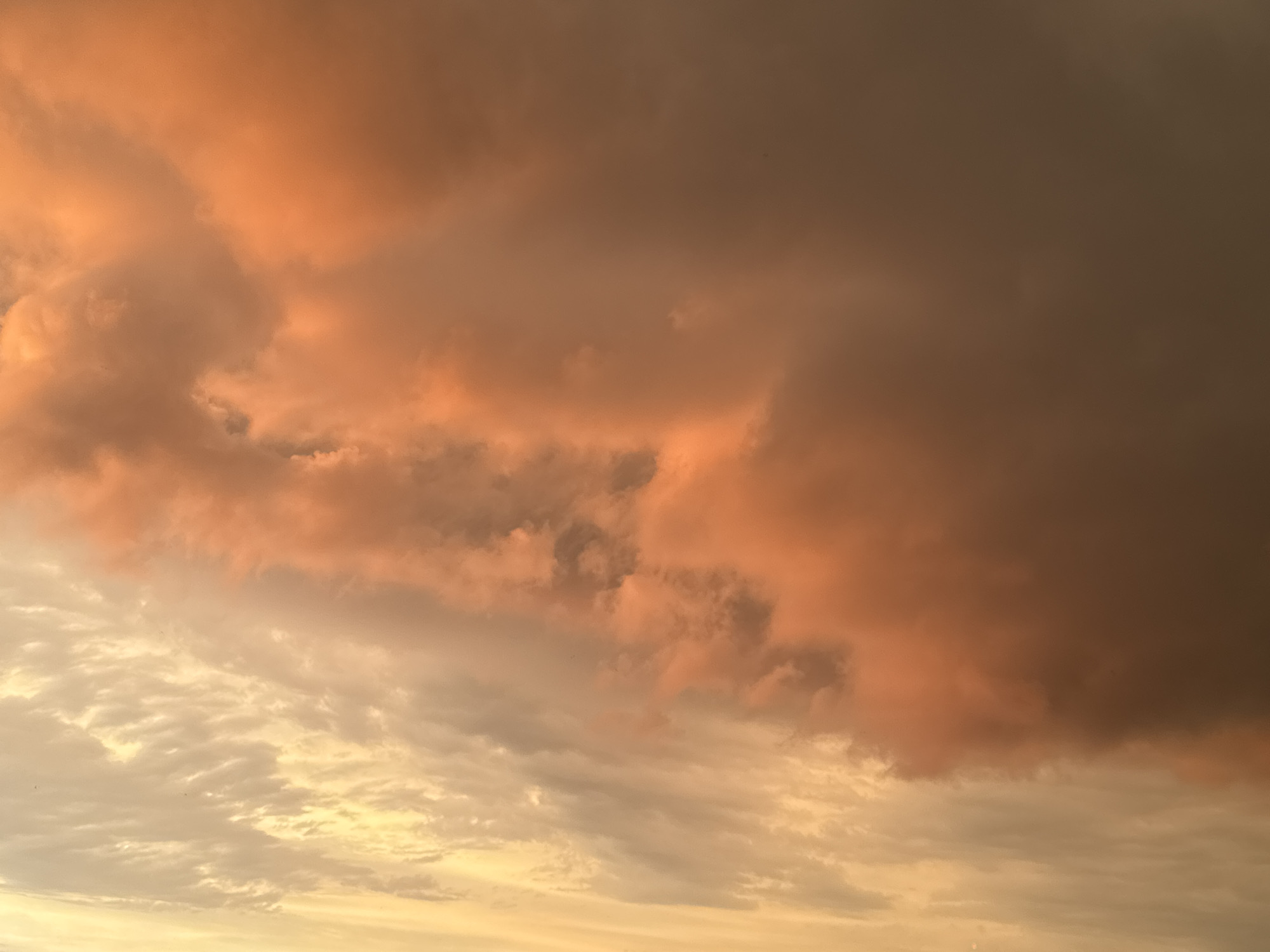 The image shows a dramatic sky filled with clouds during what appears to be sunset or sunrise. The clouds are a mix of warm oranges and darker grays. The lower portion of the sky exhibits lighter, fluffier clouds with a yellowish hue, possibly illuminated by the setting or rising sun. The upper portion is dominated by heavier, more ominous-looking clouds that are tinged with a fiery orange, suggesting they are catching the last or first rays of sunlight. The overall impression is one of a powerful and dynamic atmospheric event. There are no people, buildings, or any other discernible objects or landmarks present in the image. The setting is simply the sky.