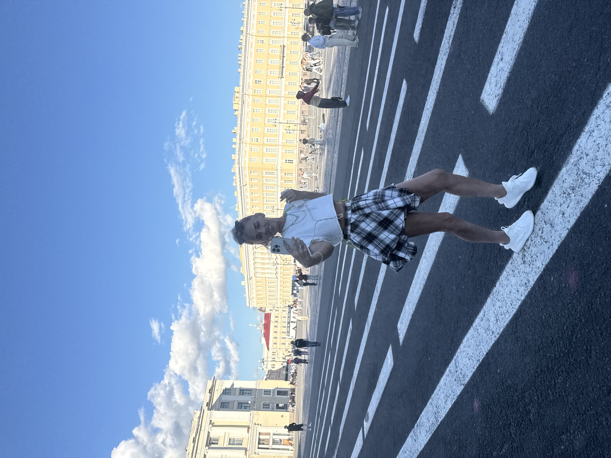 A young woman is standing on a pedestrian crossing in Palace Square, Saint Petersburg, Russia, taking a selfie with her phone. She is wearing a white crop top, a black and white plaid skirt, and white sneakers. Behind her, people are walking and gathering in the square, and large, ornate buildings, including the General Staff Building, are visible under a blue sky with scattered clouds. The sun is casting shadows, indicating it is daytime.