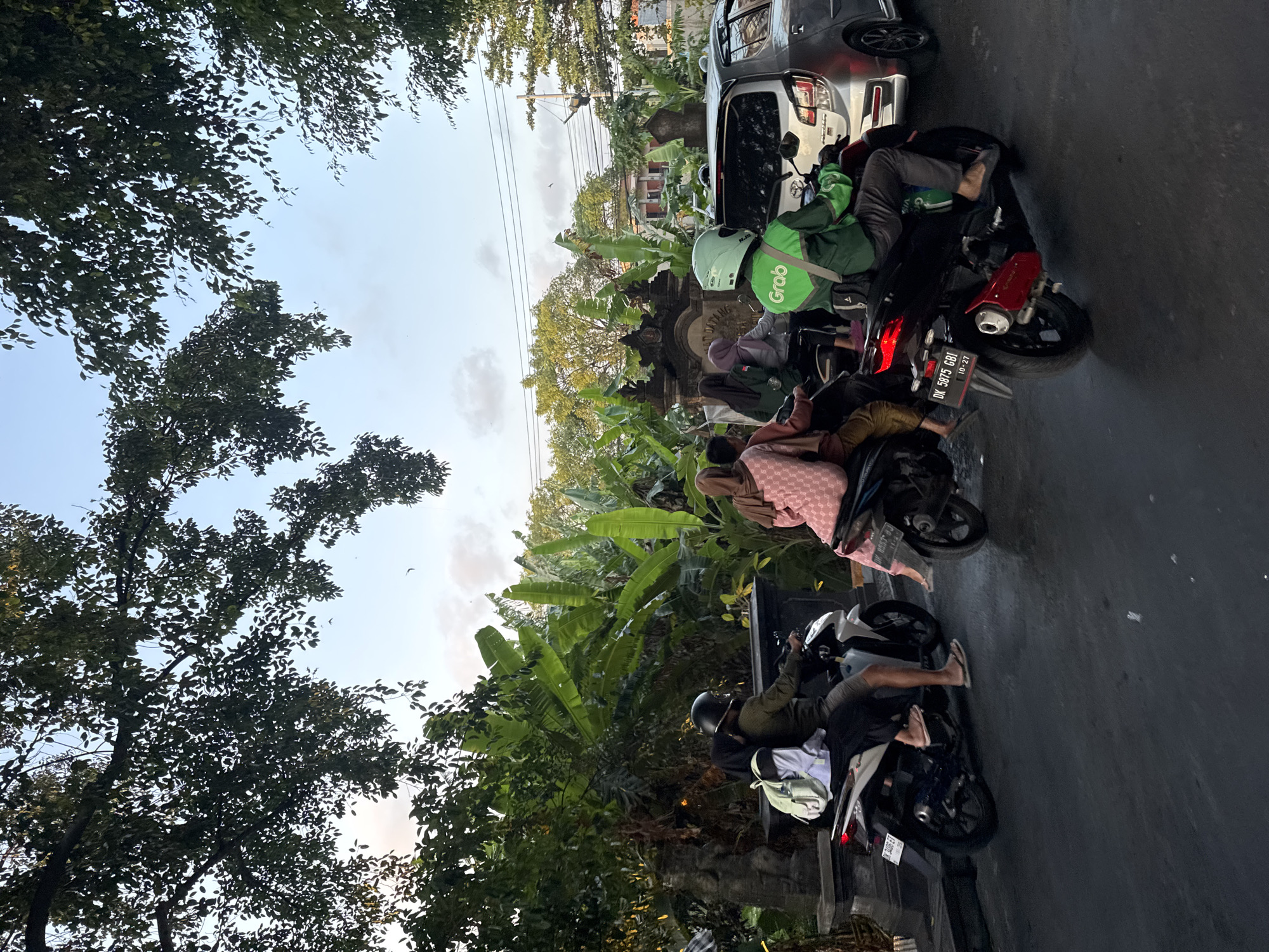 In Denpasar, Indonesia, a street scene unfolds with several people on motorcycles and a car. The foreground is dominated by a dark asphalt road, suggesting a street or thoroughfare. Lush green banana trees and foliage frame the upper and mid-sections of the image, indicating a tropical environment. In the background, a structure with ornate carvings and the word "DATANG" is visible, likely an entrance or gate to a building or complex.
Several individuals are riding motorcycles, some with passengers. One motorcycle rider, wearing a green jacket with a "Grab" logo and a light green helmet, is positioned in the mid-right of the frame, facing away from the camera and riding a red motorcycle. Another motorcycle, white, is in the mid-left, carrying two people. A third motorcycle, dark-colored, is partially visible to the right of the white one, carrying two people. A silver car is parked to the right of the Grab rider, partially obscuring the background. The sky above is a pale blue with scattered clouds, and trees with dark green leaves hang over the scene from above. The overall atmosphere appears to be a typical daytime scene in a bustling urban or suburban area of Denpasar.