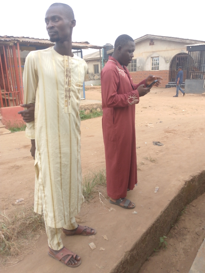 Two men stand outside, one in a beige, striped tunic with matching pants and brown sandals. The second man is in a long-sleeved, burgundy robe and sandals, looking at a cell phone. In the background, there's a street with several buildings, including one with an arched entrance and a dark metal gate. A man is walking on the street near a gray building with white-framed windows. Metal roofing structures and some scattered greenery are also visible.