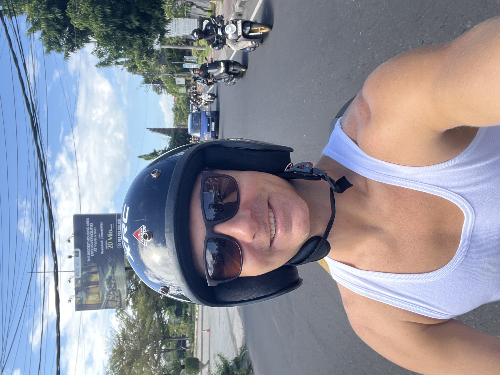 A man wearing a white tank top, sunglasses, and a black helmet is taking a selfie on a motorcycle in Denpasar, Indonesia. Several other motorcycles and a blue vehicle are in front of him on the road. A large advertising billboard for VASAKA BALI is visible to the side of the road. The billboard advertises "The Biggest Housing Area Exclusive Facilities At VASAKA BALI". It also advertises "Ready Unit", "Insentif PPN", "20 Mio", and a phone number: "081 918 350 400". Trees and a blue sky with clouds are visible in the background.