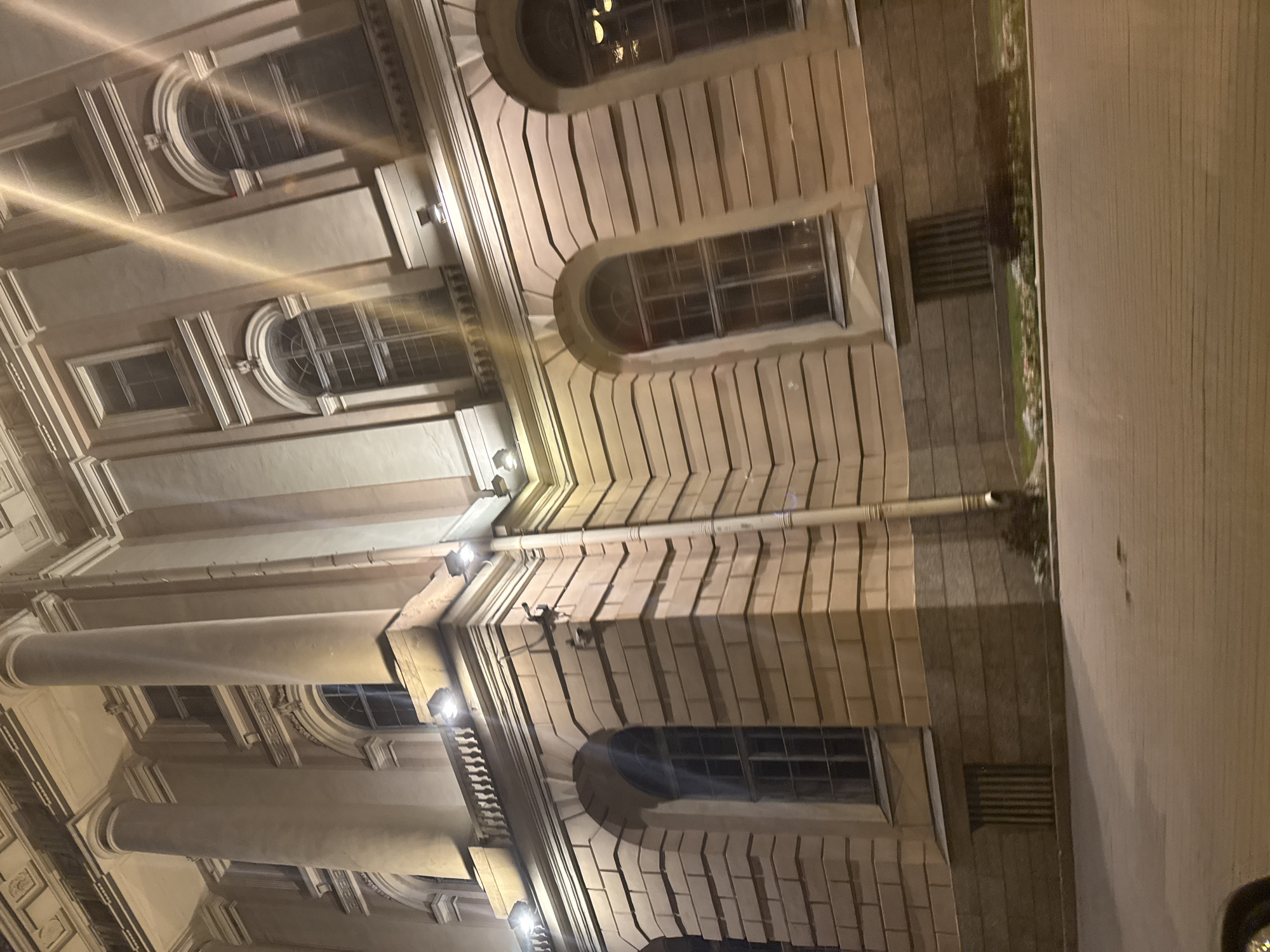 The image shows the exterior of a grand, historic building in Saint Petersburg, Russia, at night. The architecture is classical, with large, fluted columns supporting an entablature. The facade is constructed of light-colored stone blocks, many of which are arranged in horizontal courses. There are several arched windows with metal grates, and some windows are rectangular. The building is illuminated by several modern, rectangular lights mounted on the facade. A downspout runs vertically along the side of the building. In the foreground, a paved walkway or street is visible, with some low-lying greenery and flower beds bordering the building. The overall impression is one of imposing, old-world architecture enhanced by contemporary lighting.