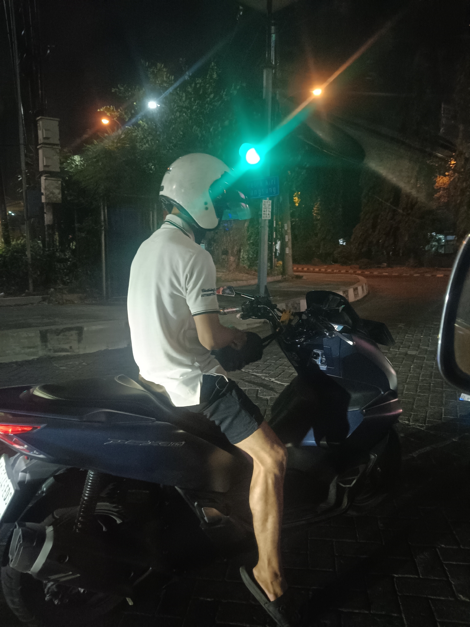 A person wearing a white helmet and a white polo shirt sits on a dark-colored scooter at night. The person is wearing black shorts and appears to be at a traffic light. The traffic light is green. The scene takes place on a street paved with dark cobblestones, with trees and a building visible in the background. The lighting suggests it is nighttime, possibly in an urban area. The image does not provide enough context to definitively determine the location as Surabaya, Indonesia.