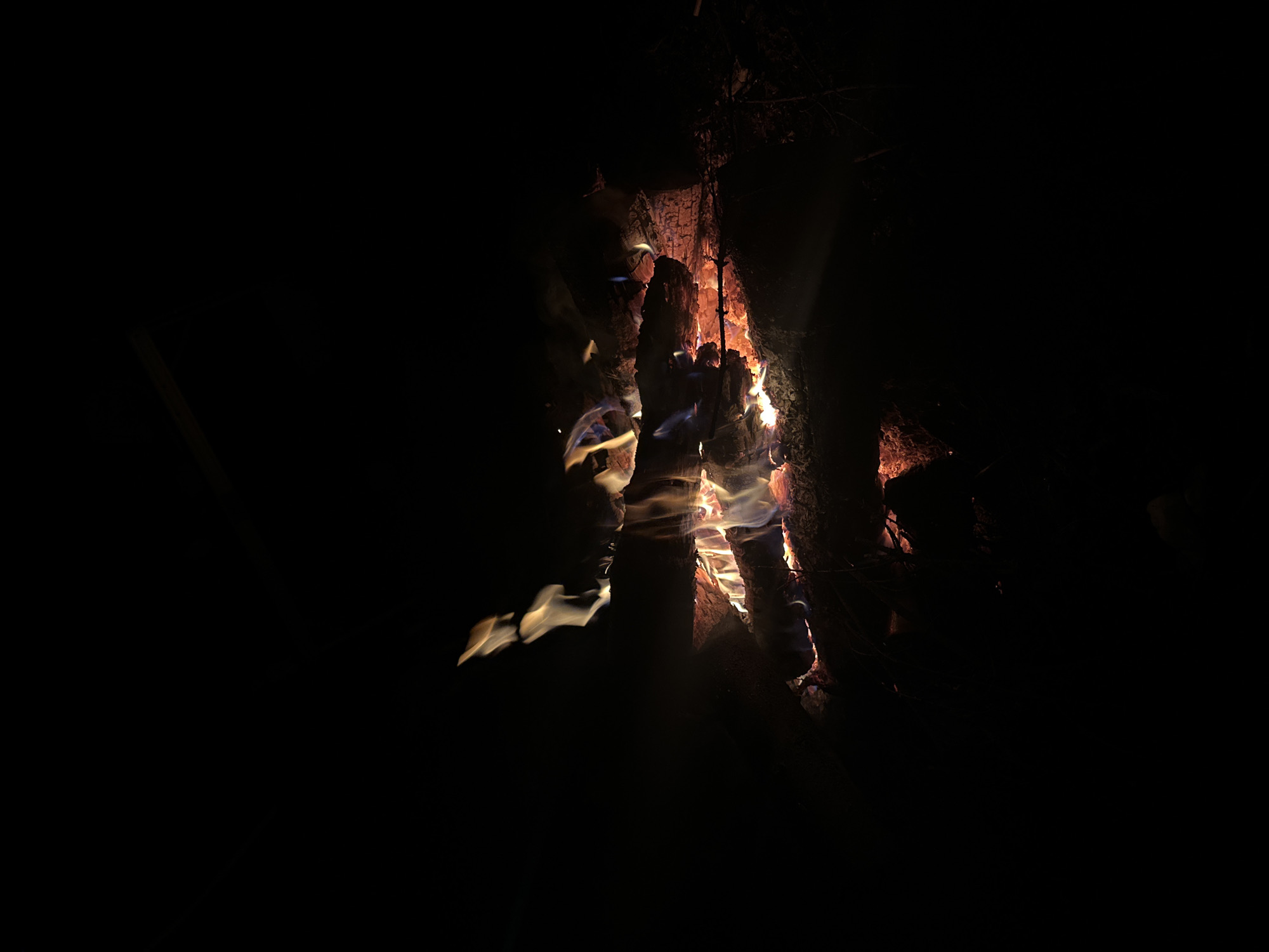 A campfire burns in the dark in Папярнянскі сельскі Савет, Belarus. Logs are stacked vertically and horizontally, with flames licking around them. The surrounding area is shrouded in darkness.