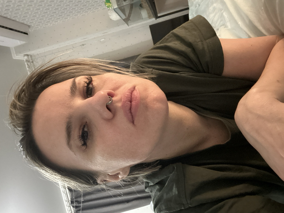An adult female is depicted in a close-up, head-and-torso shot, positioned indoors in what is identified as Rostov-on-Don, Russia. The individual has light brown or blonde hair, styled away from her face, and appears to be looking directly into the camera. She has a silver-colored ring piercing in her nasal septum. Her eyes show long, dark eyelashes, and her lips are slightly parted. She is wearing a dark olive-green short-sleeved t-shirt. Her left forearm is visible in the lower right portion of the frame, suggesting a reclining or supported posture. The background on the left displays dark grey drapery. On the right, a white air conditioning unit is mounted high on a wall adorned with light-colored, patterned wallpaper. Below the air conditioning unit, a glass enclosure, resembling a terrarium, is visible on a surface, with a white, indistinct object positioned nearby.